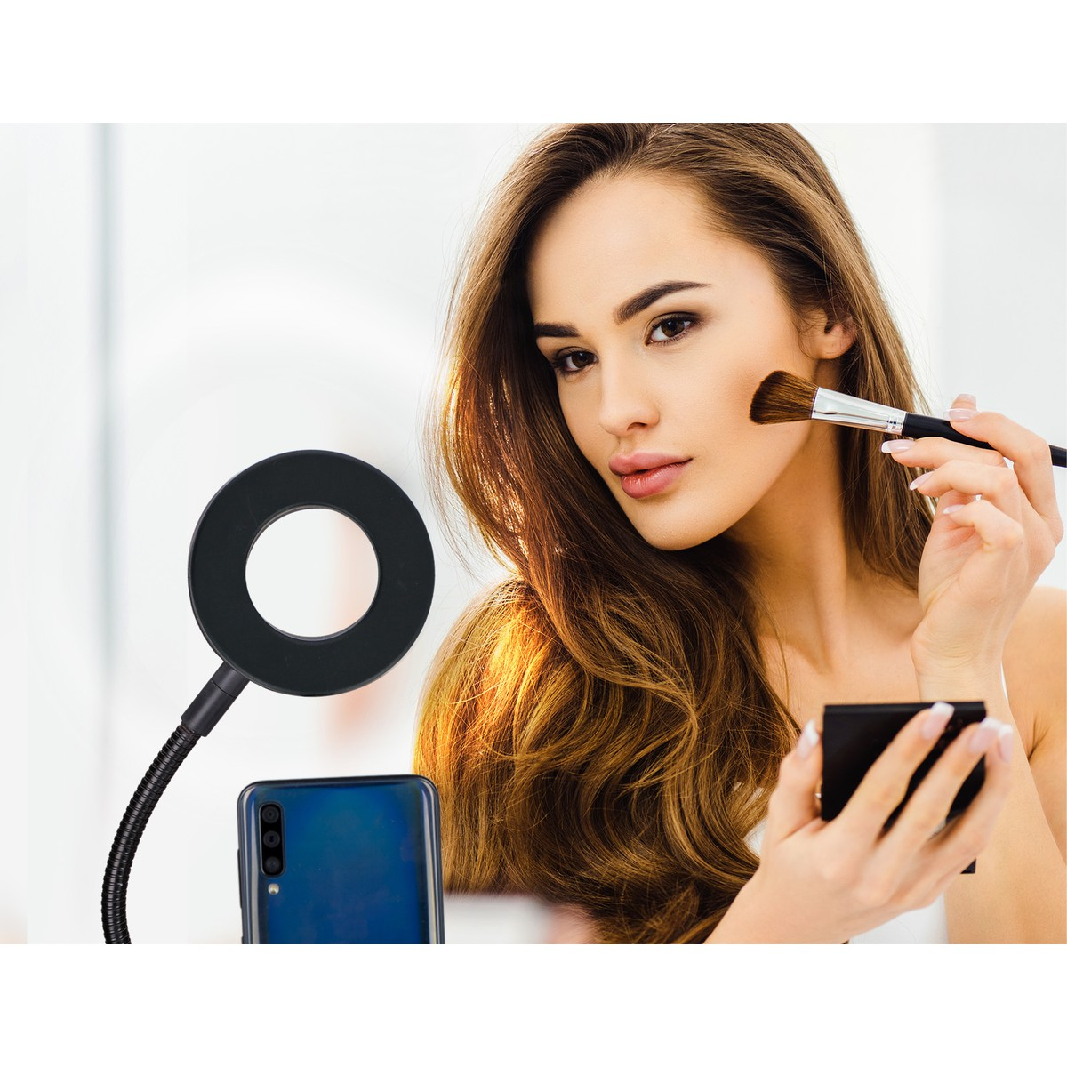 Woman applying makeup with a brush and mirror, with a ring light and phone nearby.