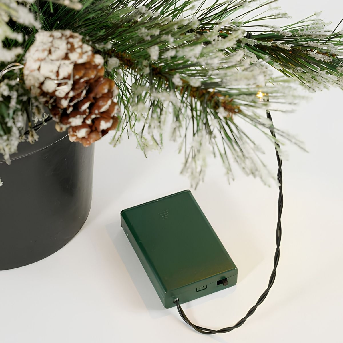 Green battery box with string lights next to a pot of pine branches and pinecones.