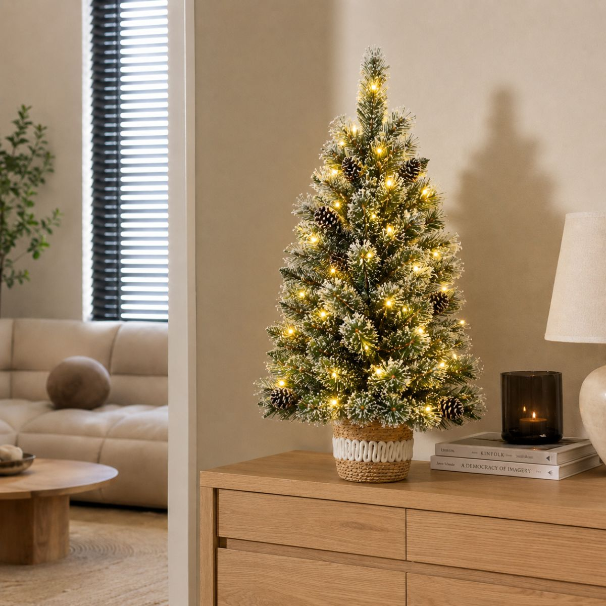 A small, frosted Christmas tree with pinecones and lights sits on a wooden dresser.