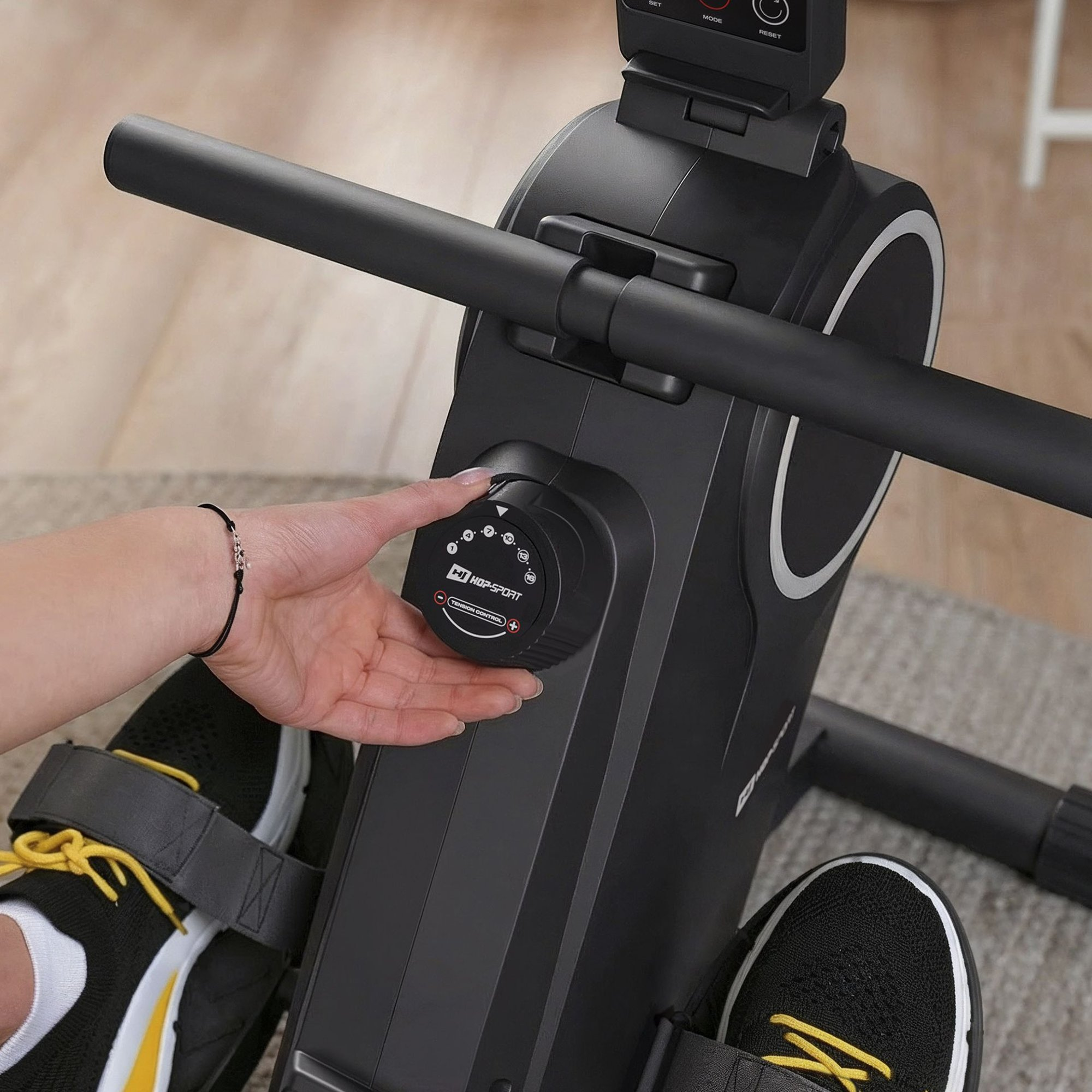Eine Hand dreht den Widerstandsregler einer schwarzen Rudermaschine, während Füße in Sportschuhen auf den Pedalen ruhen.