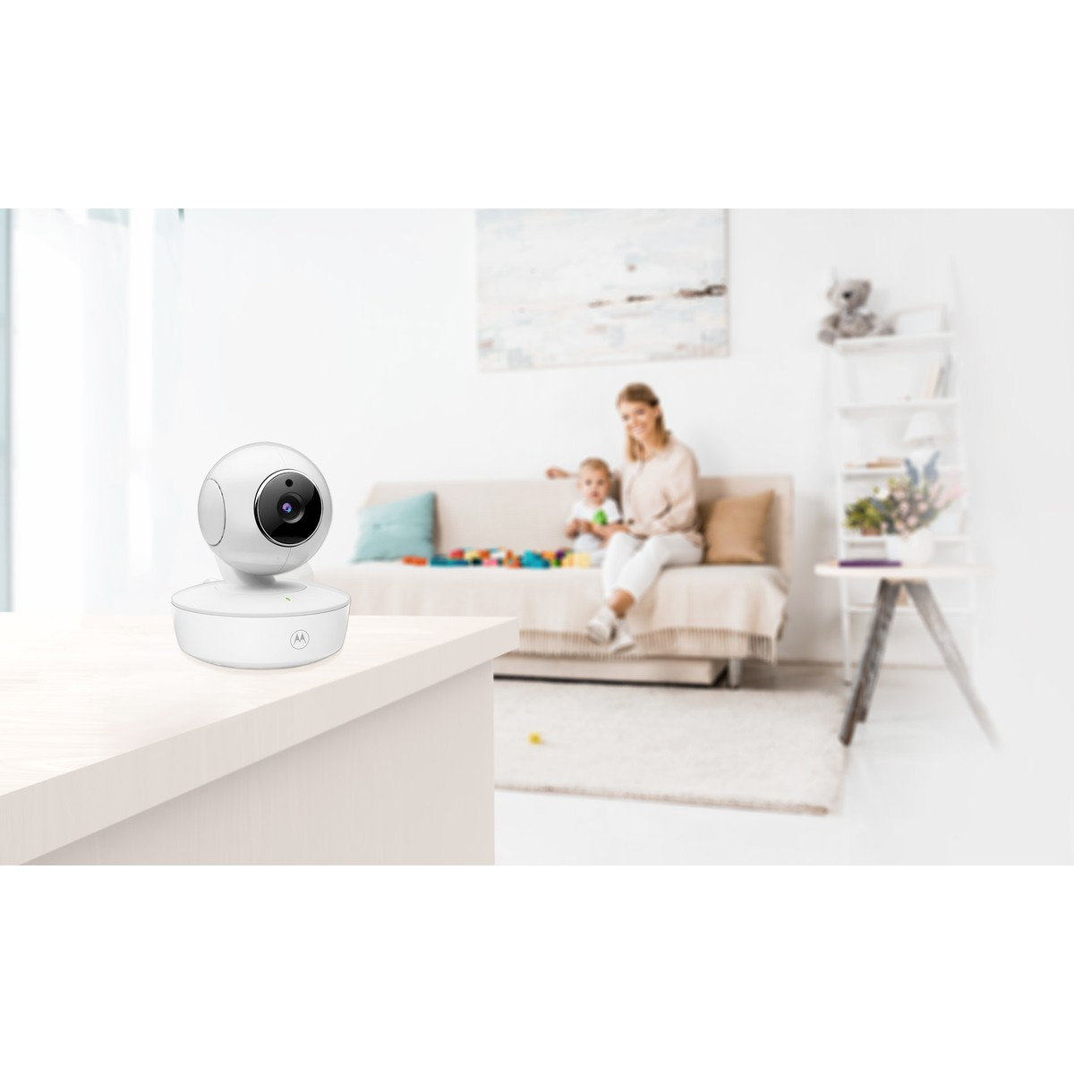 A white baby monitor sits on a table. A woman and child sit on a sofa in the background.