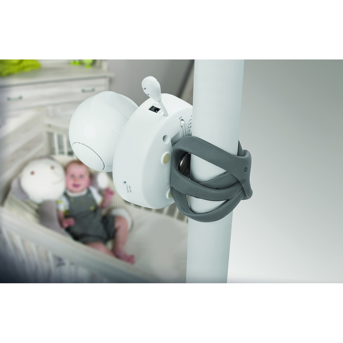 A baby monitor with camera attached to a pole, with a baby in the background.