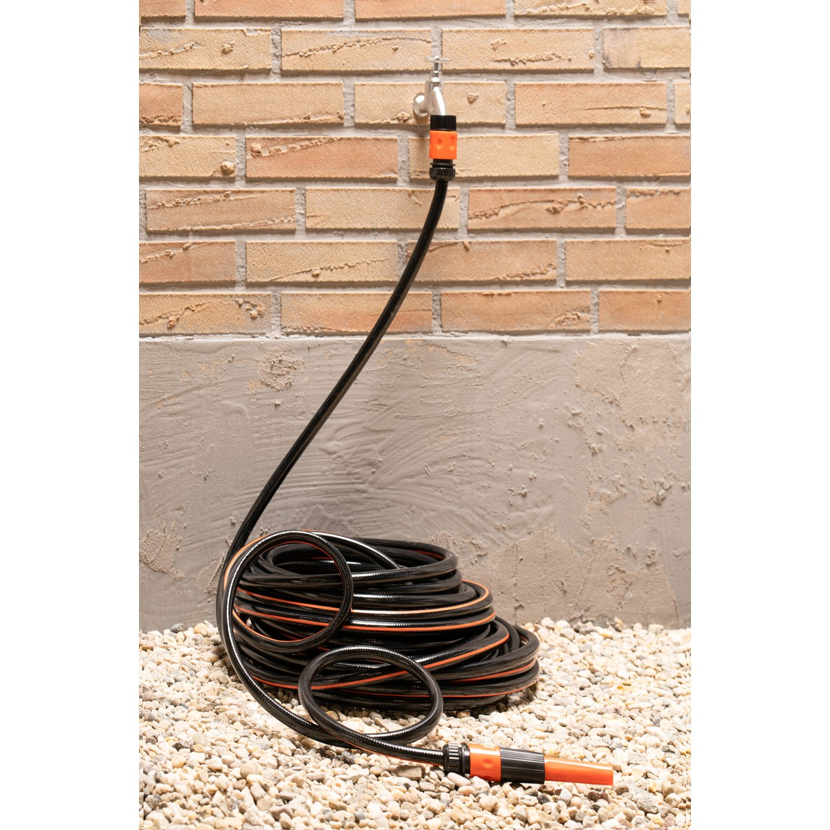 Black garden hose with orange accents, coiled on gravel against a brick wall.