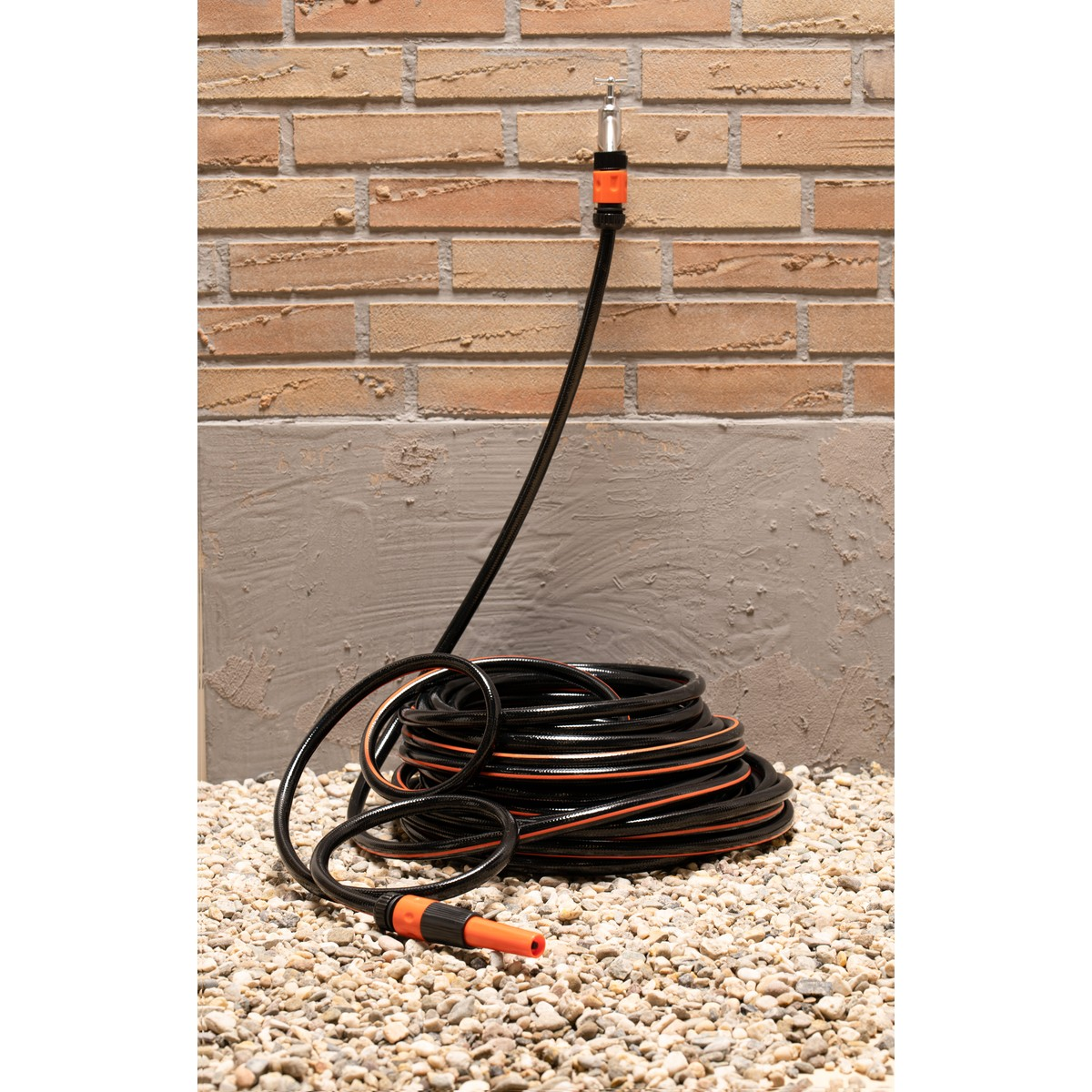 Black garden hose with orange accents on gravel in front of a brick wall.