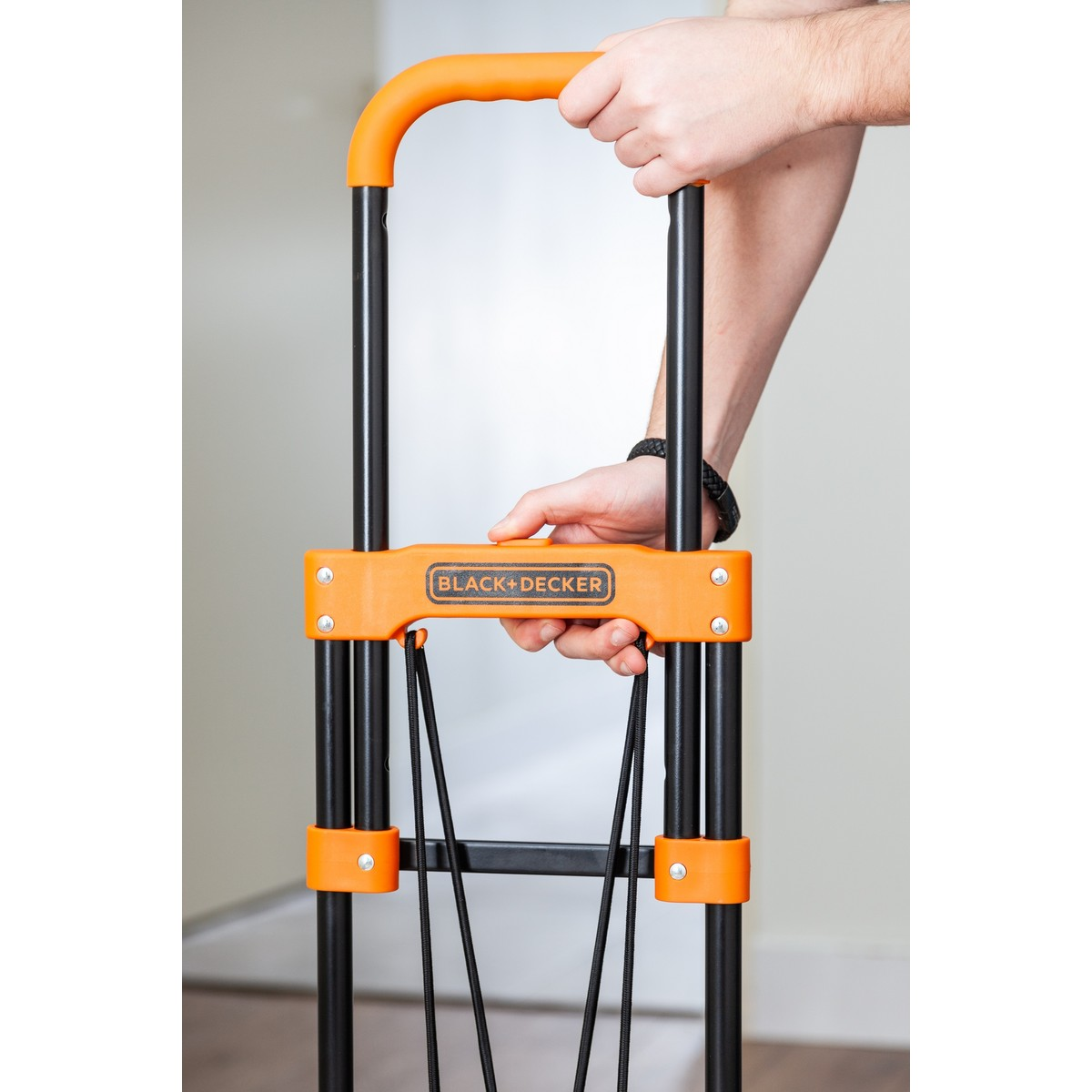 Person holds an orange folding hand truck with black handles and elastic cords.