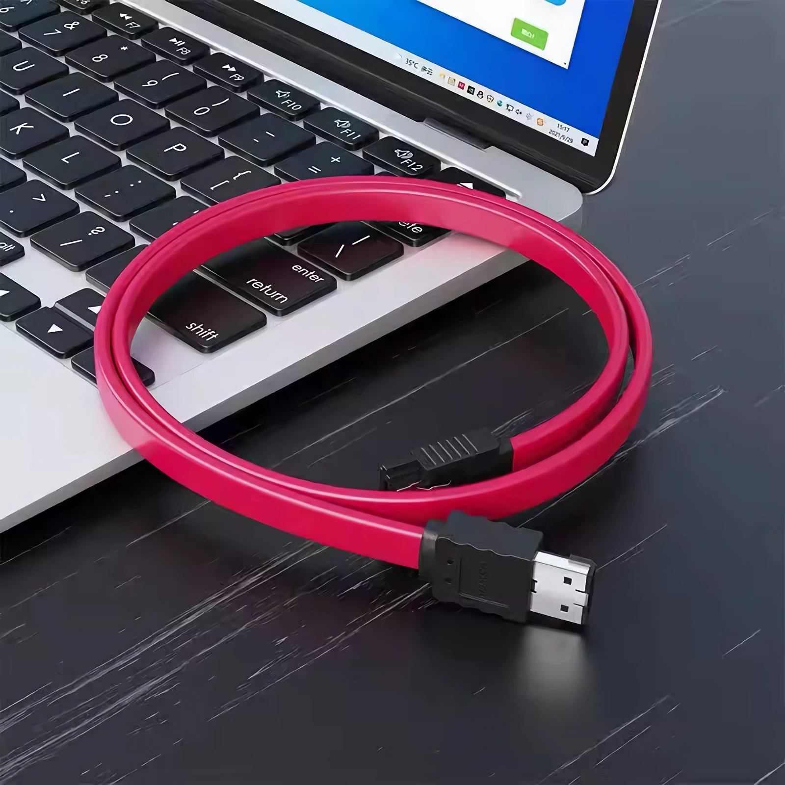Ein leuchtend pinkes SATA-Kabel mit schwarzen Anschlüssen liegt auf einer silbernen Laptop-Tastatur, mit einem Bildschirm, der eine blaue Oberfläche anzeigt.
