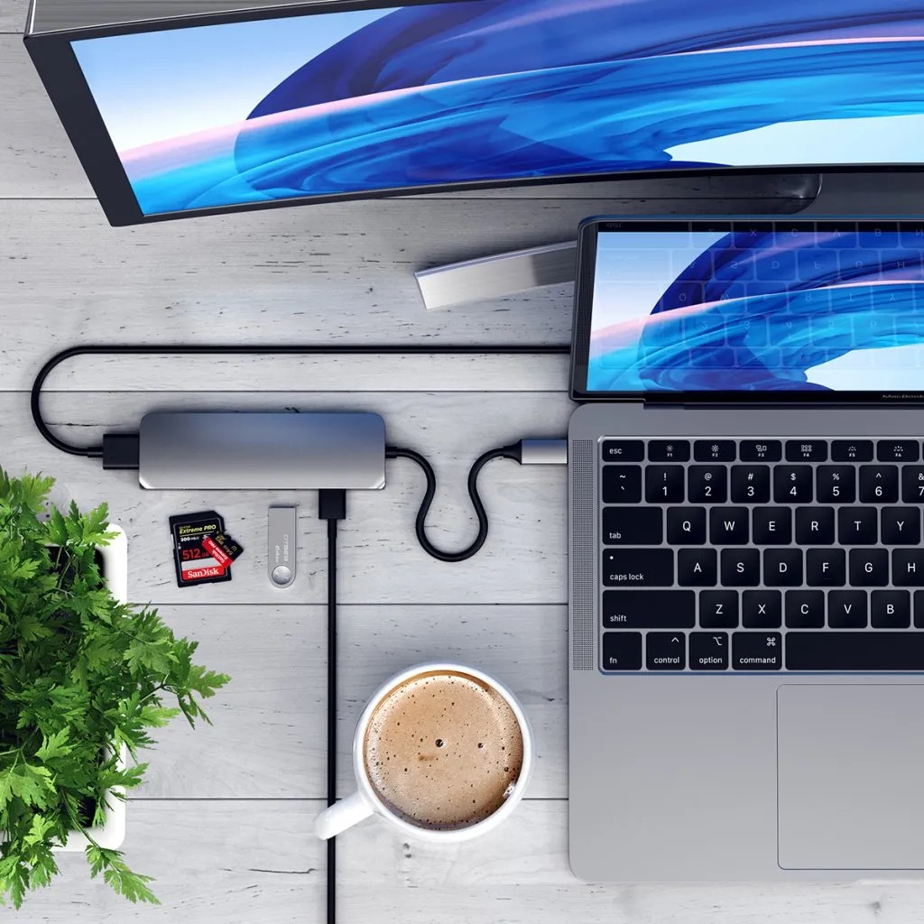 Laptop, monitor, hub, karty SD, pendrive i kubek kawy na drewnianym stole.