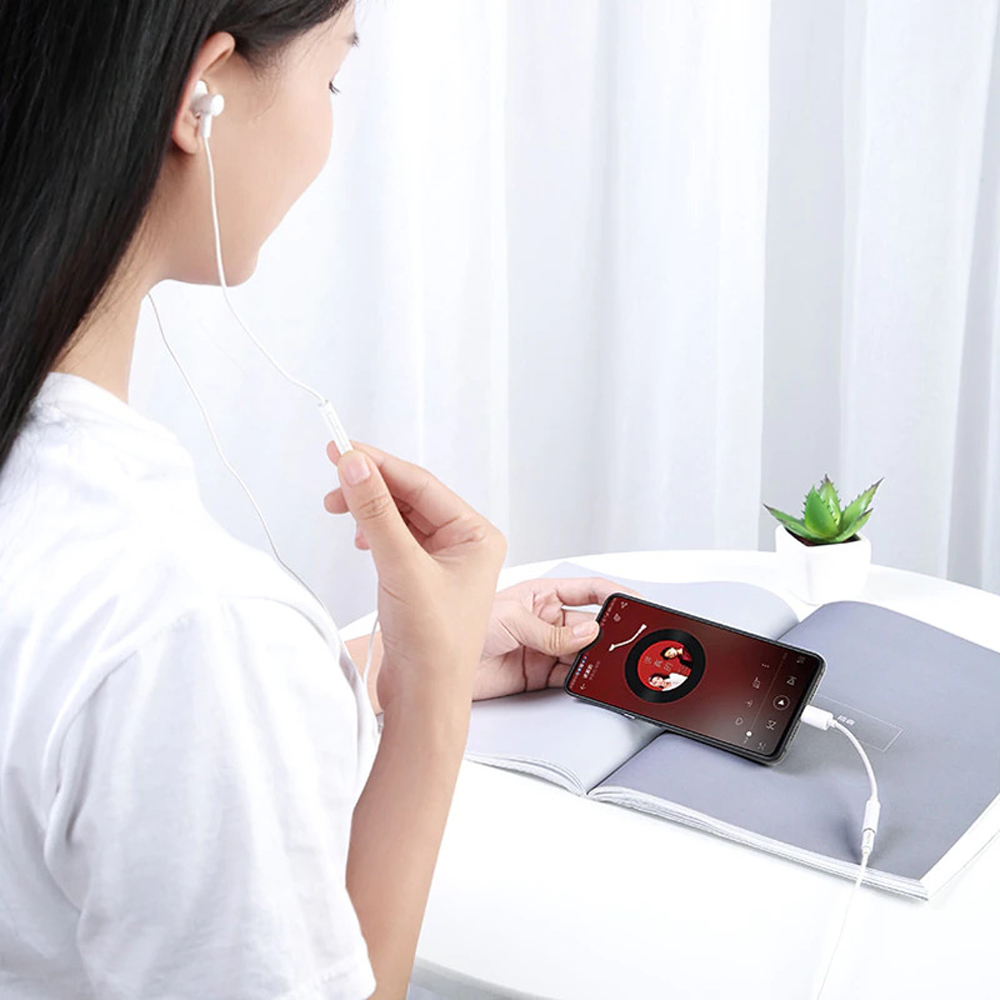 Kadın müzik çalan telefona beyaz kulaklık bağlıyor. Yakında küçük bir bitki var.