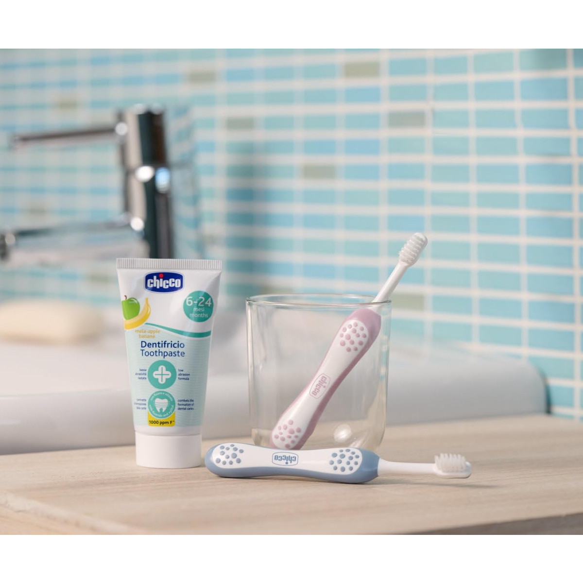 Tubo di dentifricio Chicco e due spazzolini, uno blu, uno rosa, in un bicchiere.