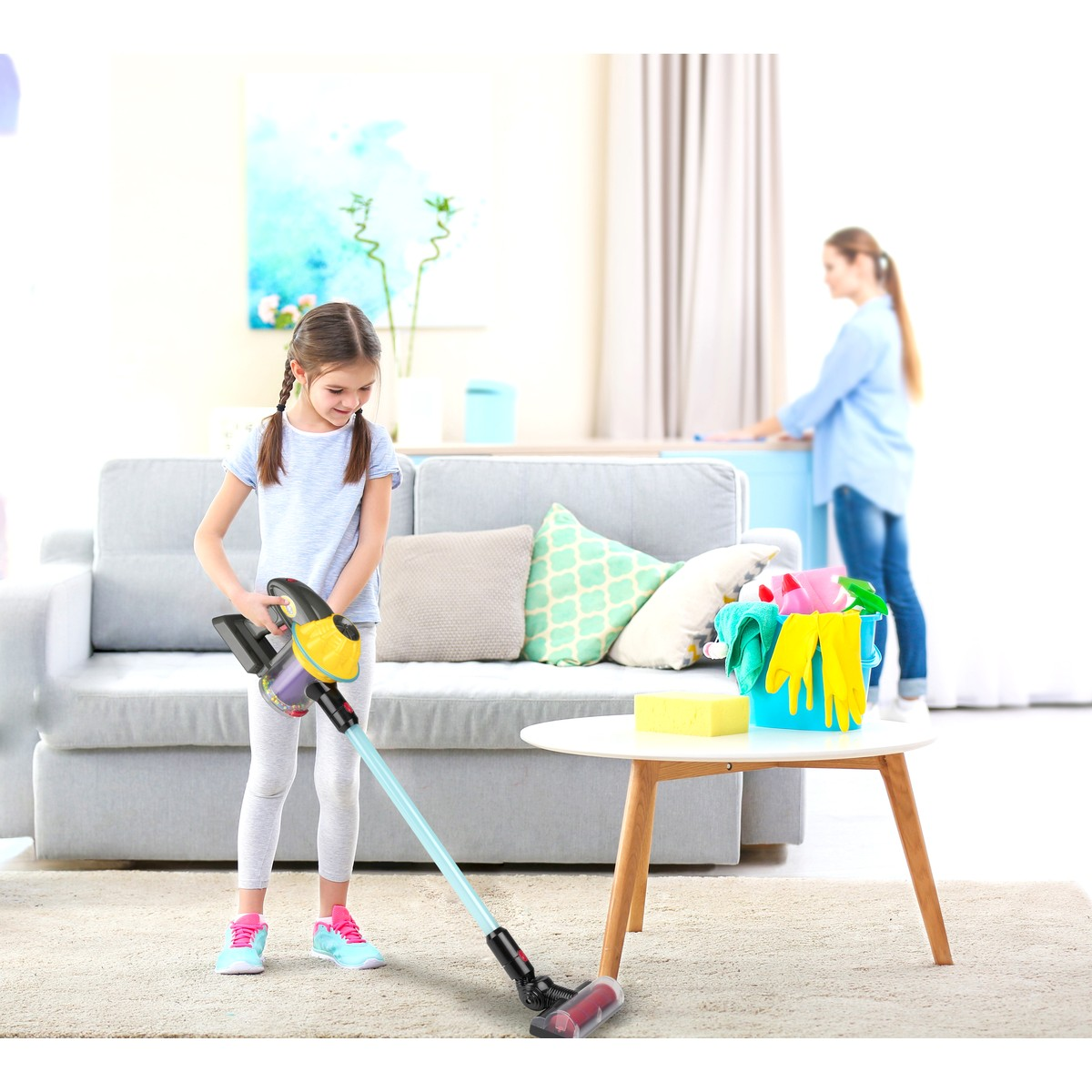 A girl vacuums with a toy vacuum, a woman in the background.