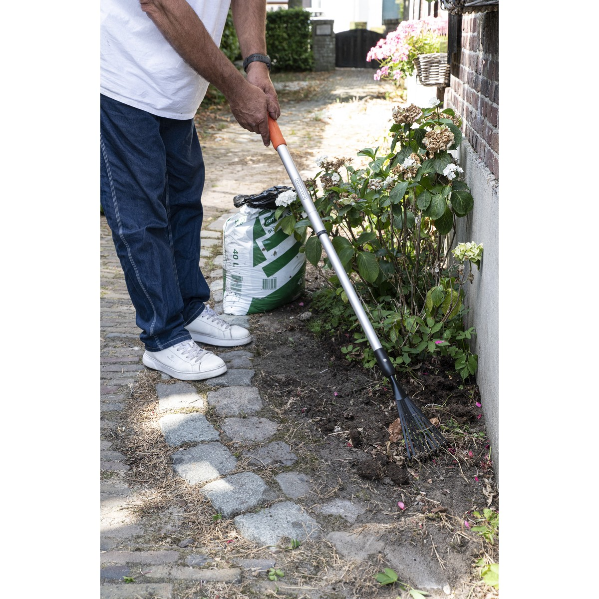 Man with rake in garden next to bag of soil and a flowerbed.