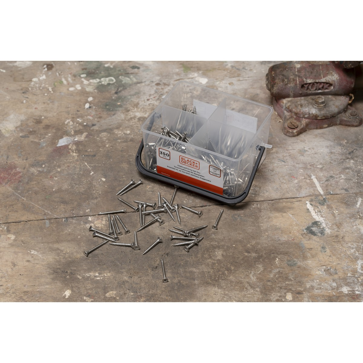 A clear plastic container holds many screws, with some scattered on a wooden surface.
