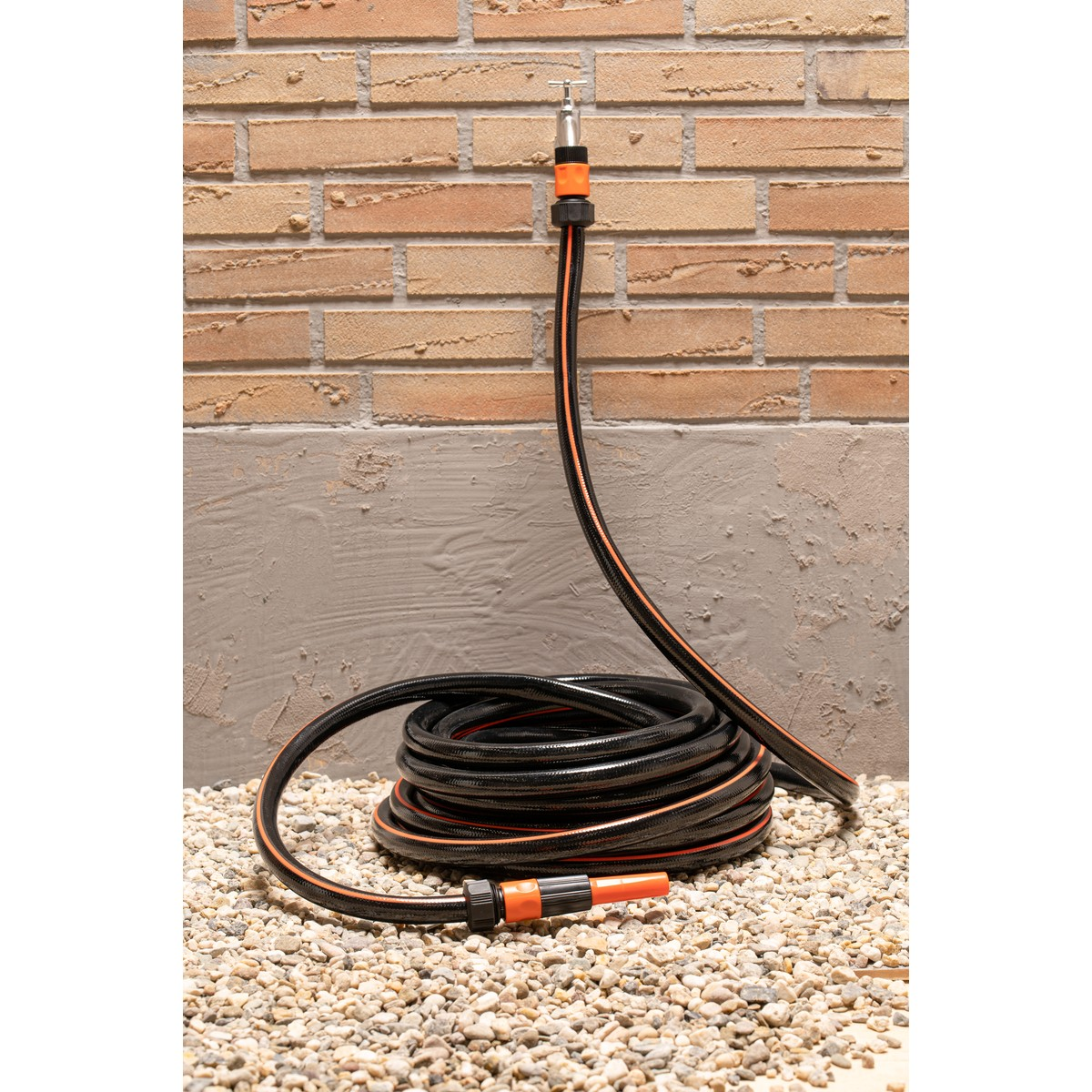 A black garden hose with orange trim is coiled on gravel in front of a brick wall.