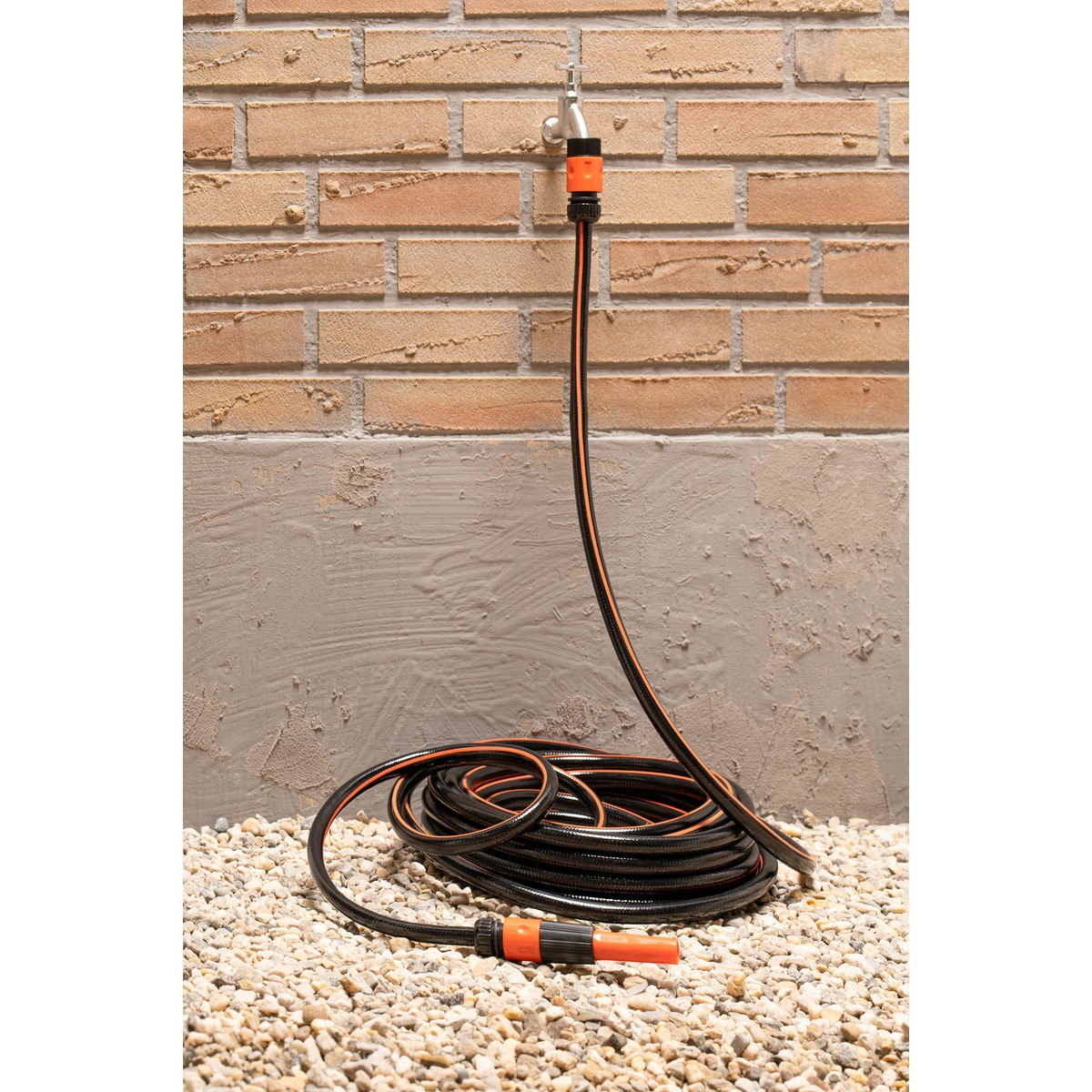 Black garden hose with orange connectors, coiled on gravel, against a brick wall.