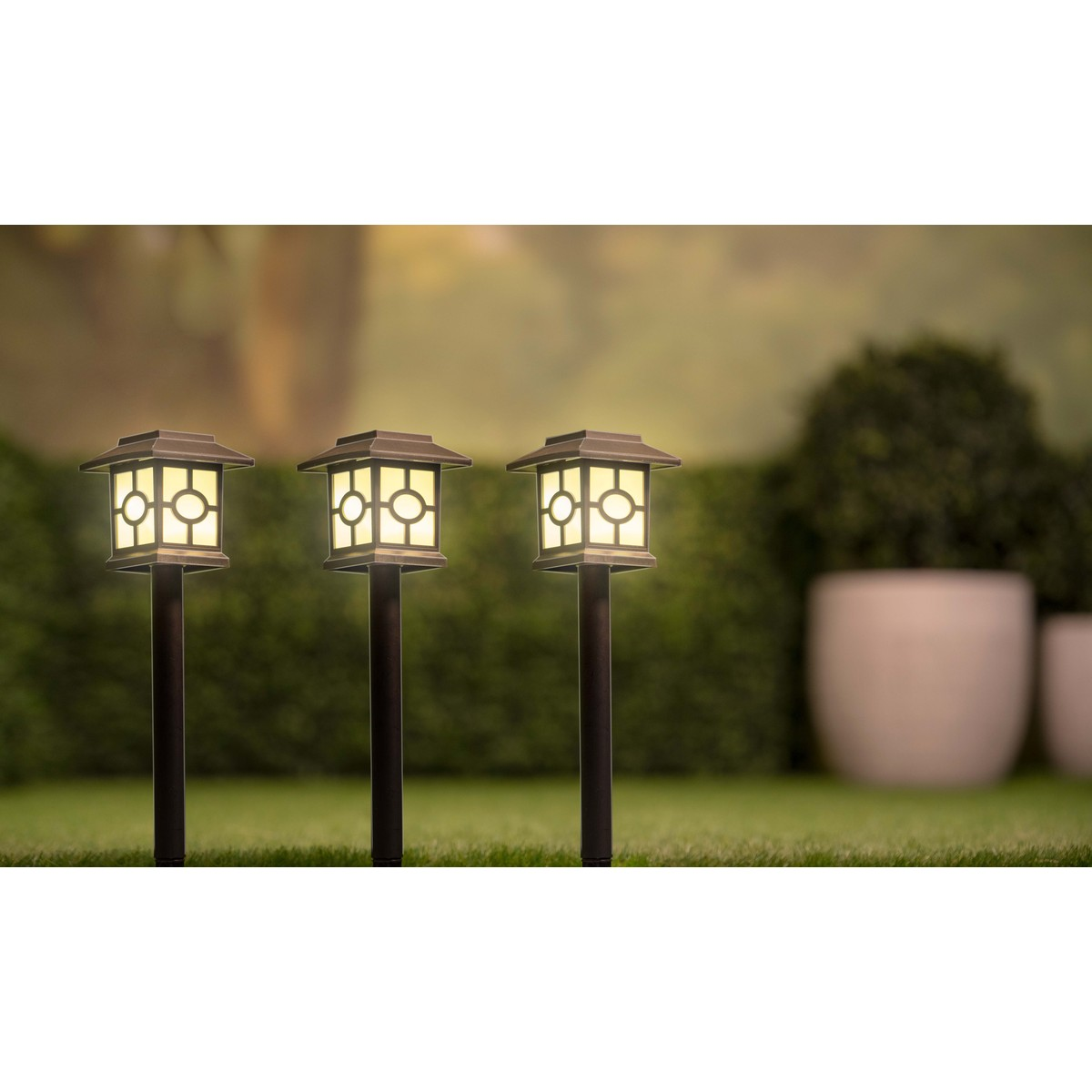Three dark path lights with geometric glass on a lawn, with blurred bushes and planters.