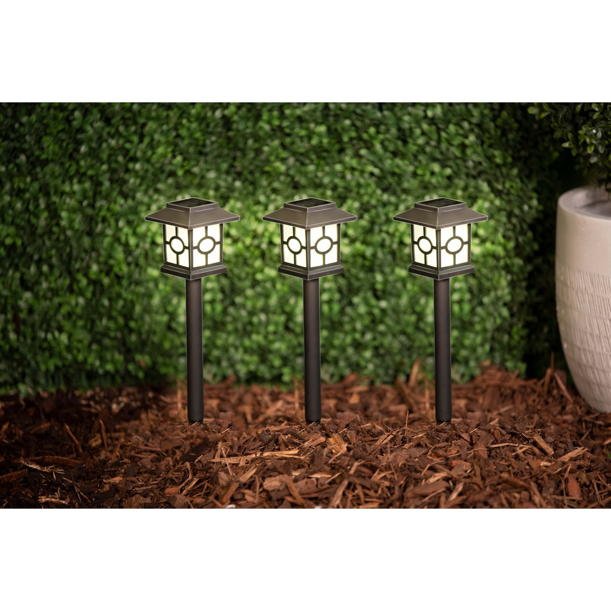 Three dark pathway lights with square lanterns stand in mulch before a green hedge.
