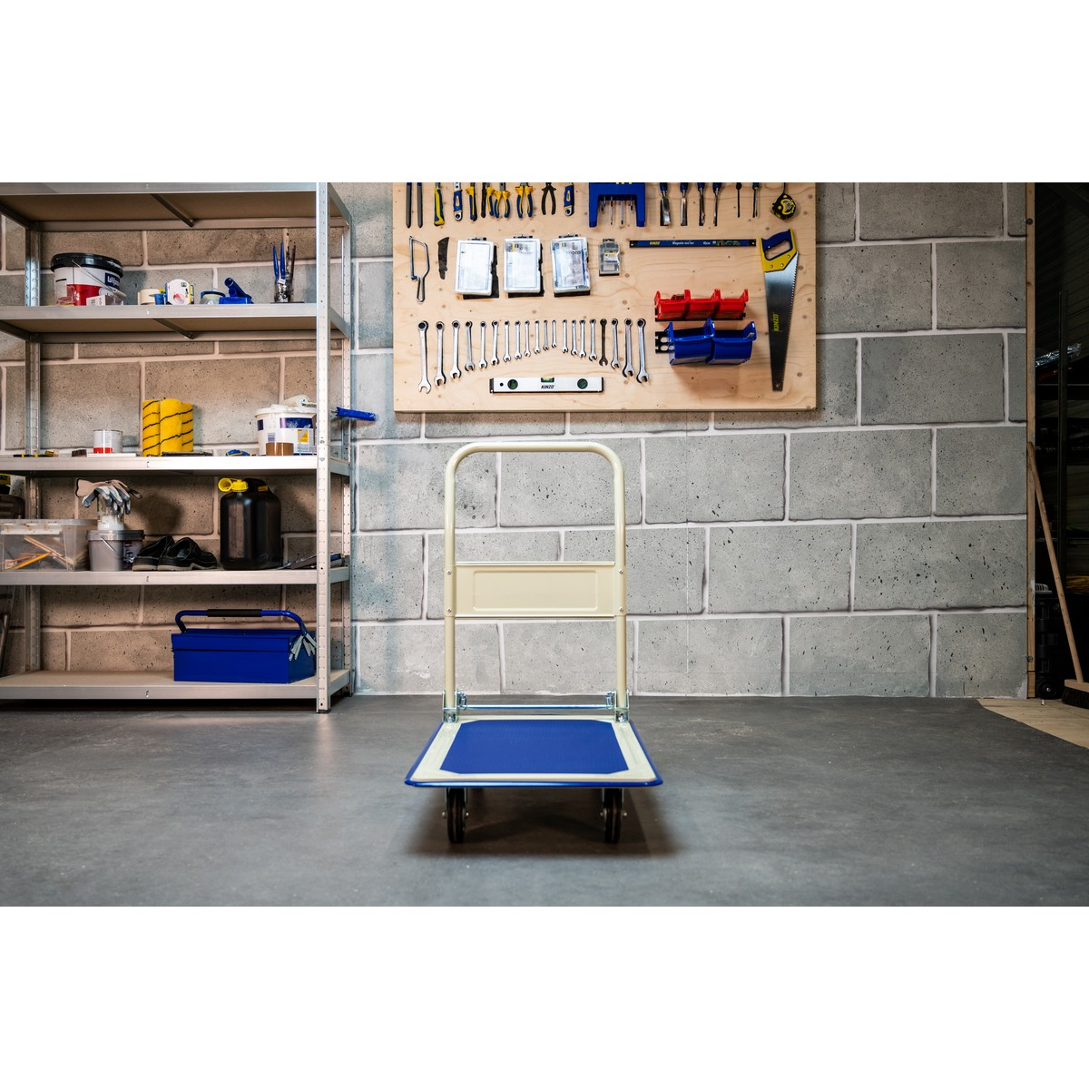A hand truck with blue and beige handle stands before a tool wall and shelves.