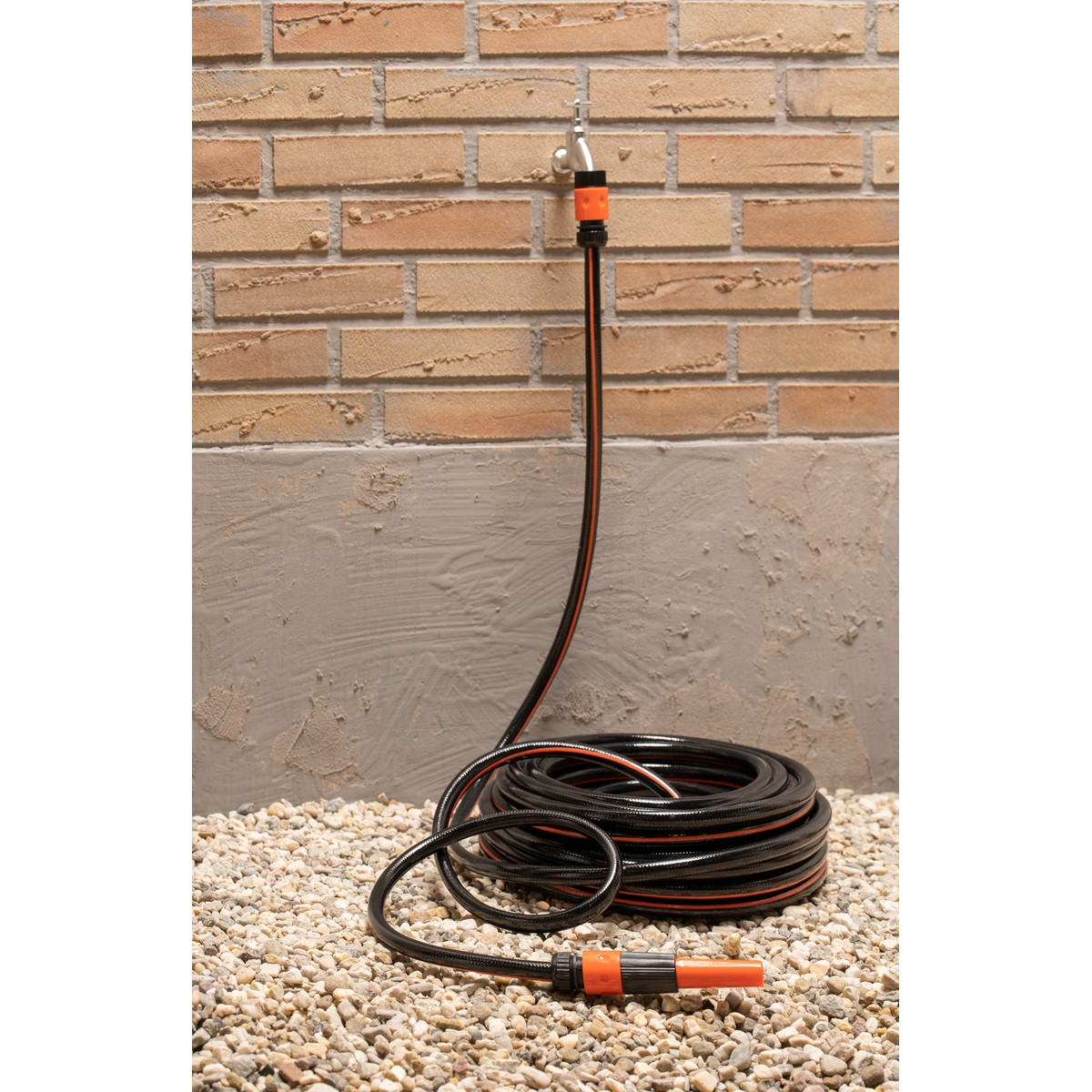 Black garden hose with orange stripes, coiled on gravel in front of a brick wall.