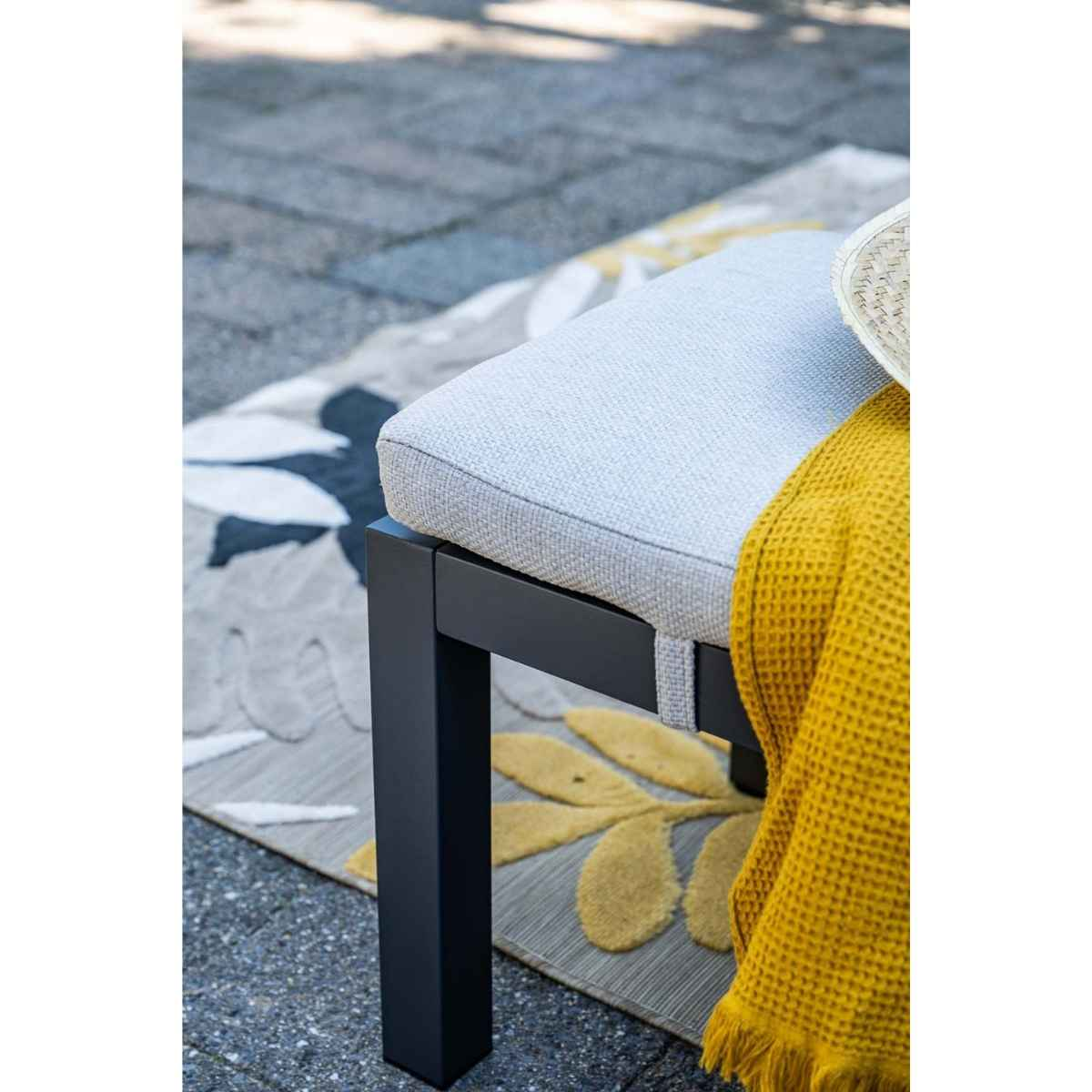 A light gray bench with a yellow blanket and hat on a floral patterned rug.