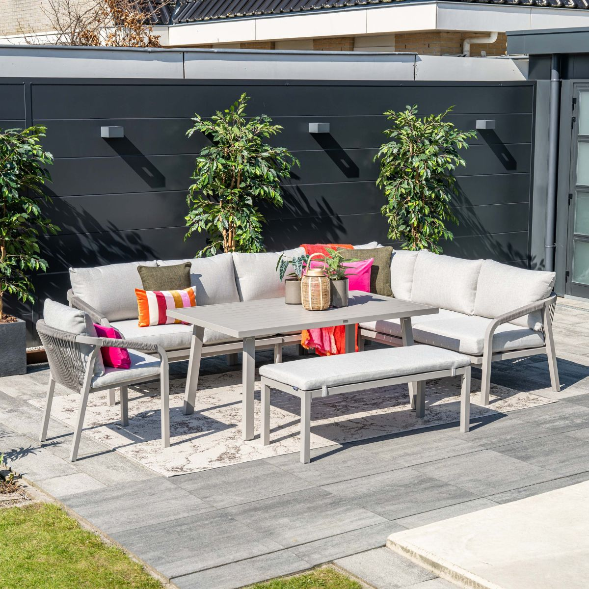 Gray outdoor lounge set with table and bench on a paved patio, surrounded by plants.