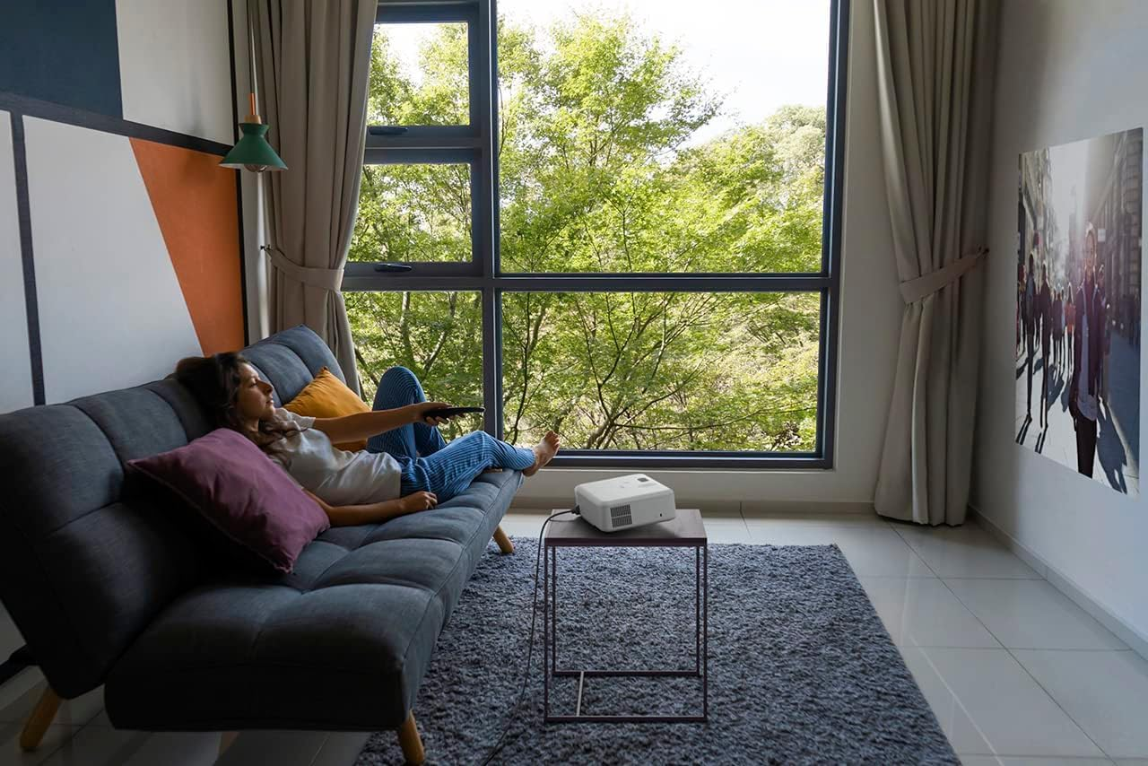 A woman relaxes on a sofa, watching a projector. Trees are visible outside.