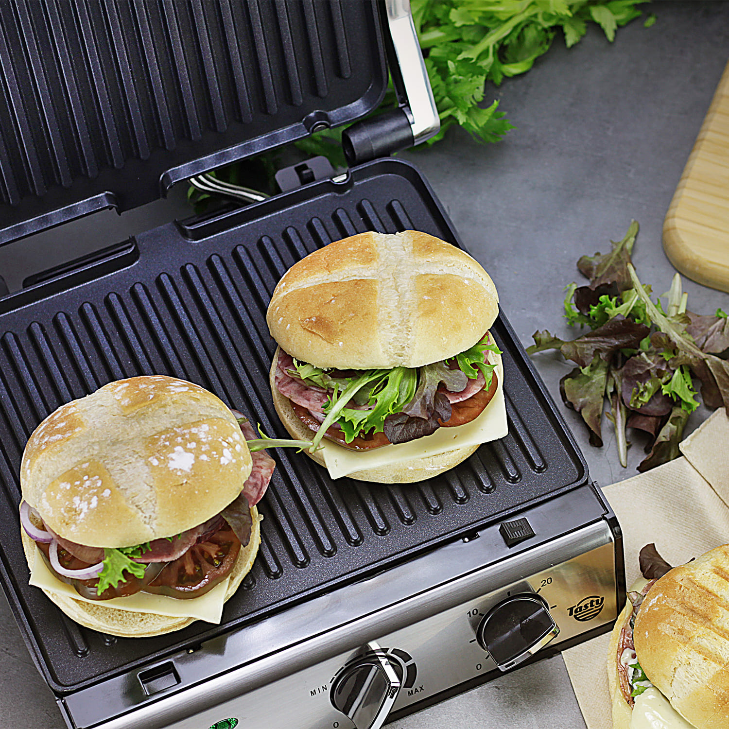 Dos sándwiches en una parrilla, con ensalada y tabla de cortar al fondo.