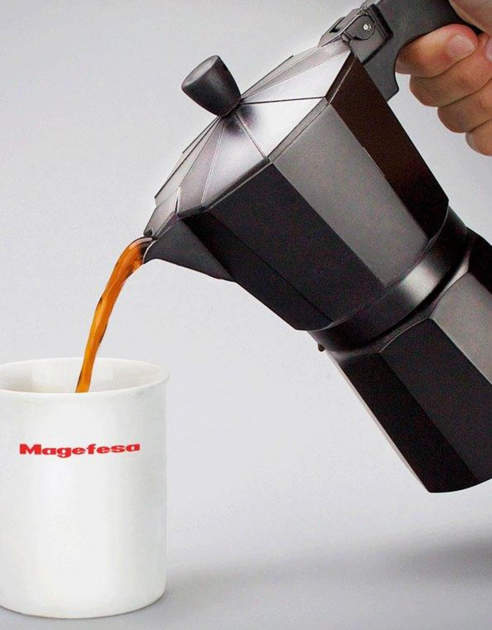 Una cafetera moka negra vierte café en una taza blanca con logo Magefesa.