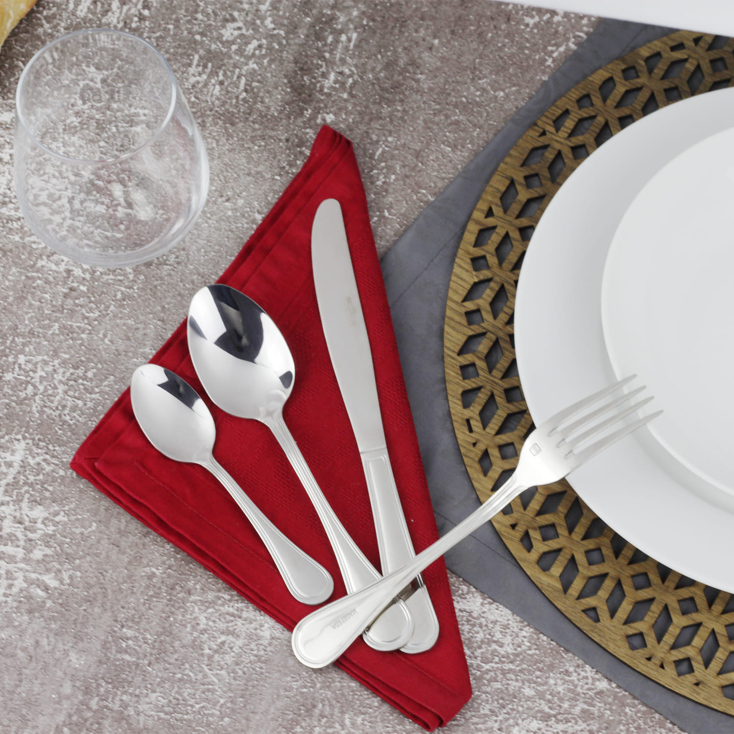 Una mesa puesta con cubiertos sobre servilleta roja, vaso y platos sobre mantel individual.