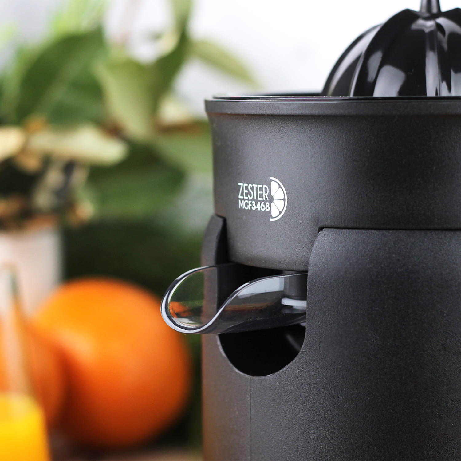 Un exprimidor de cítricos Zester MCF3468 negro con caño para jugo de naranja. Hay naranjas y hojas verdes detrás.