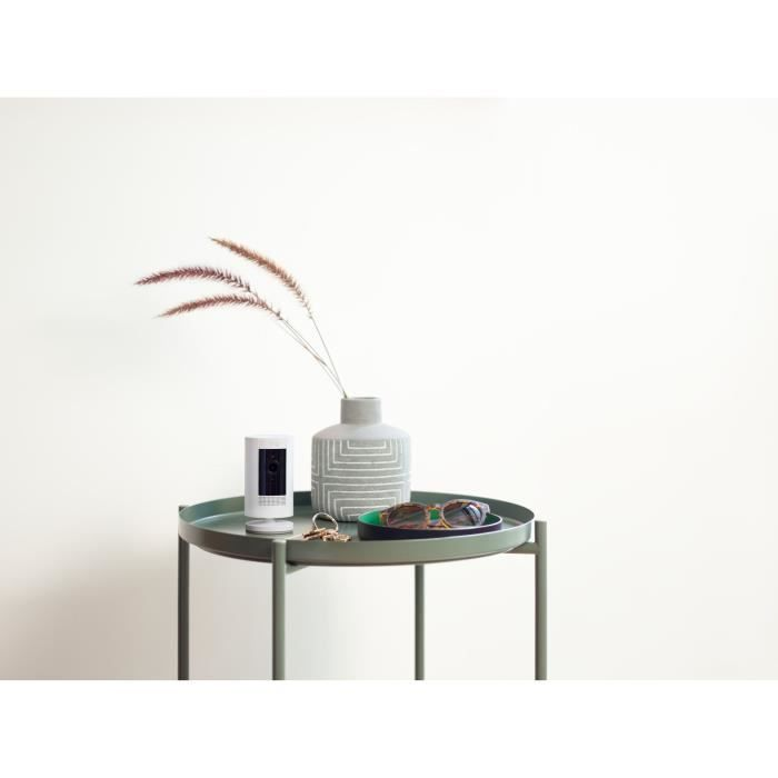 Una telecamera di sicurezza bianca, chiavi, occhiali da sole e un vaso con erba secca su un tavolo verde.