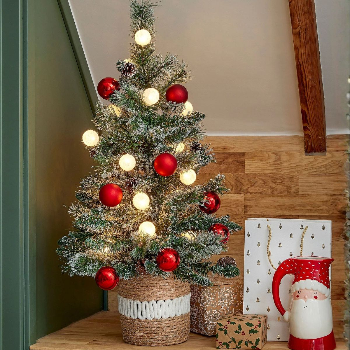 Een kleine kerstboom met rode ballen en lampjes, naast cadeaus en een kerstman mok.