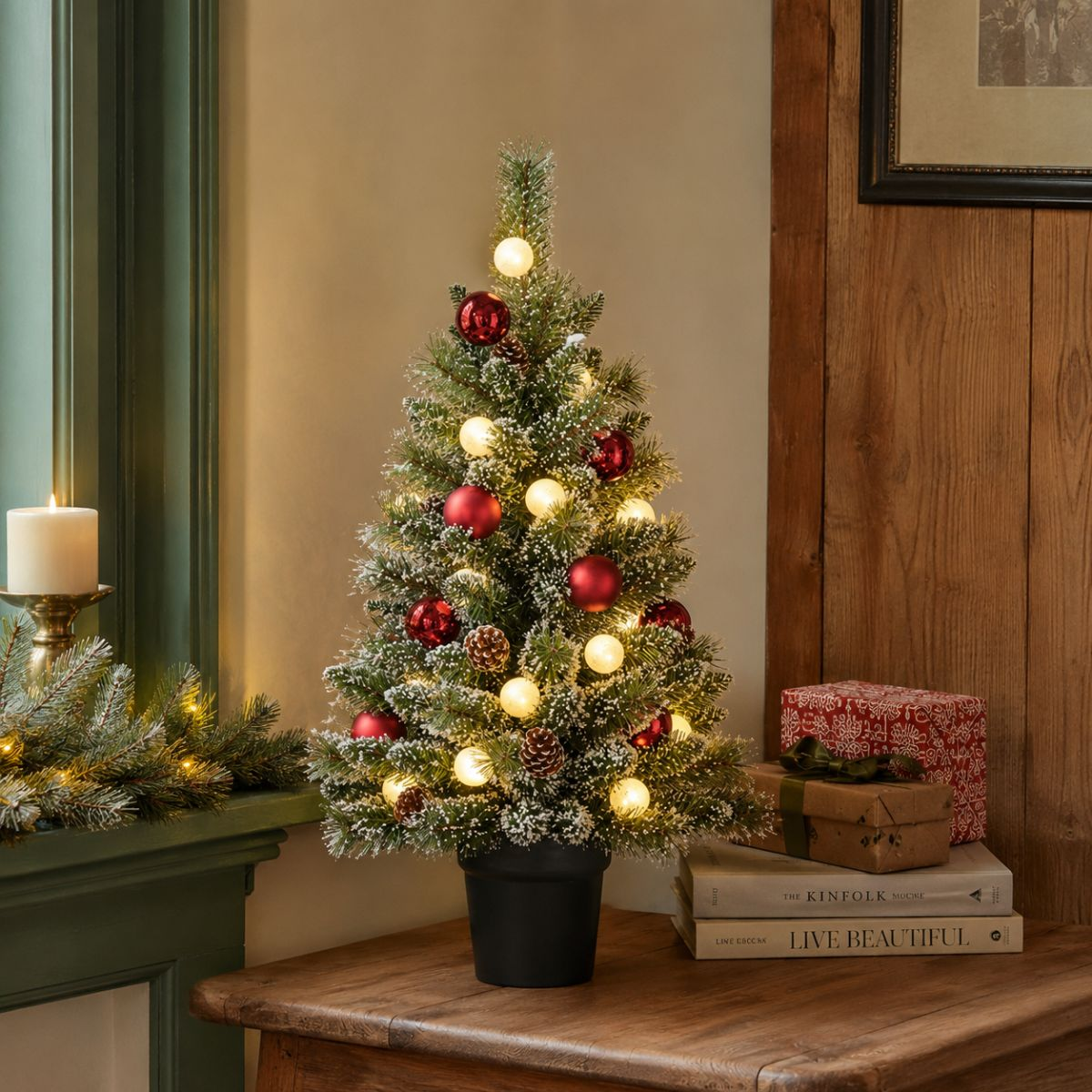 Een kleine, besneeuwde kerstboom met rode ballen en lampjes staat op een tafel.