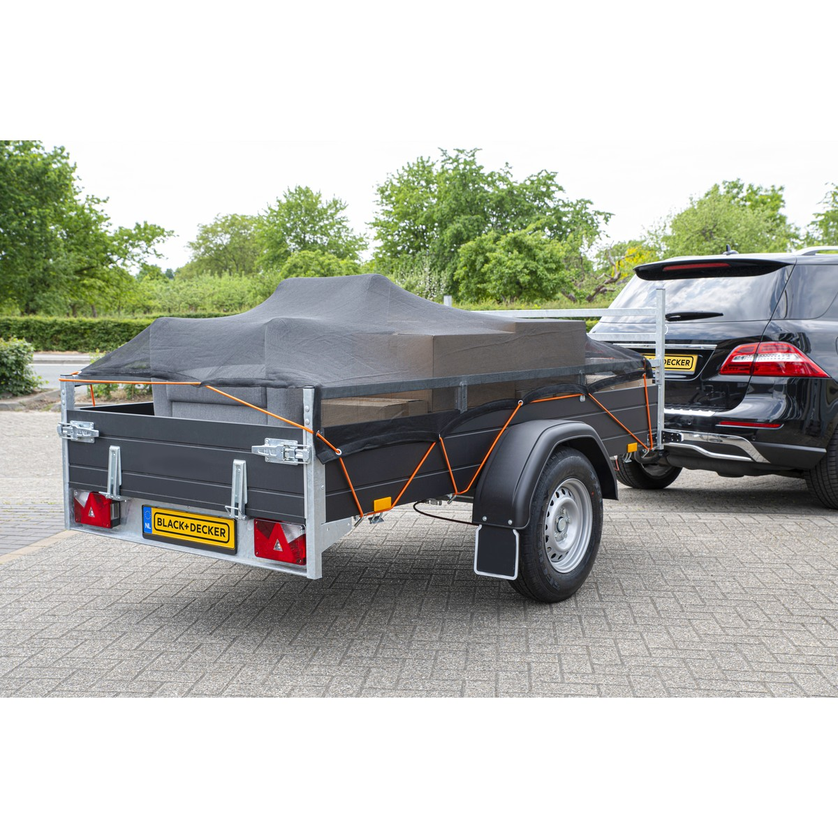 Zwarte aanhanger met lading, vastgezet met net en touwen, achter een zwarte auto.
