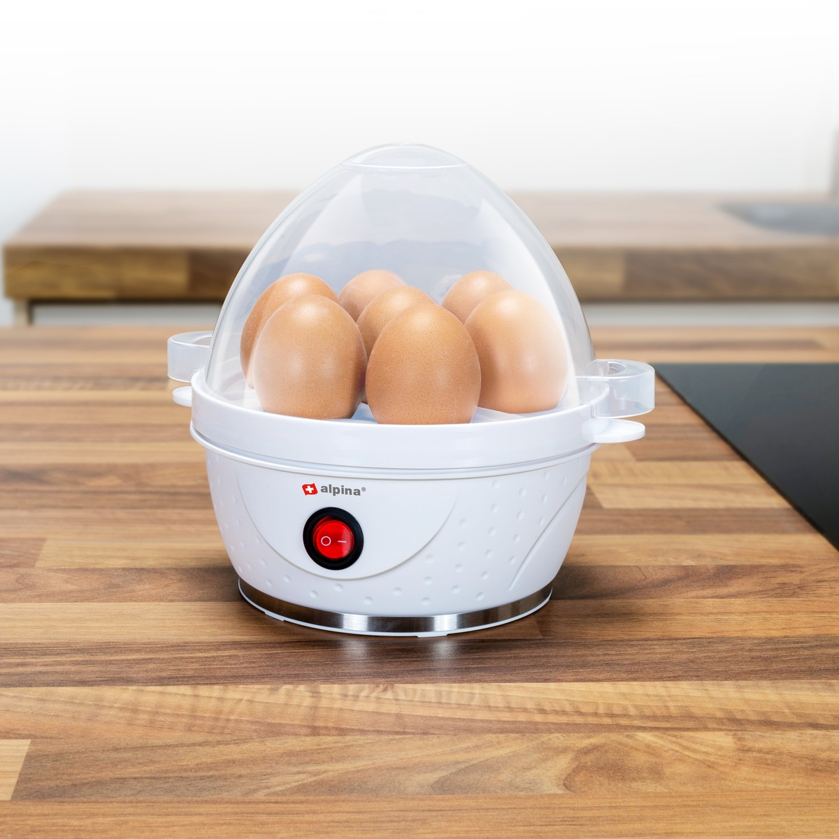 A white egg cooker with six brown eggs under a clear lid on a wooden counter.