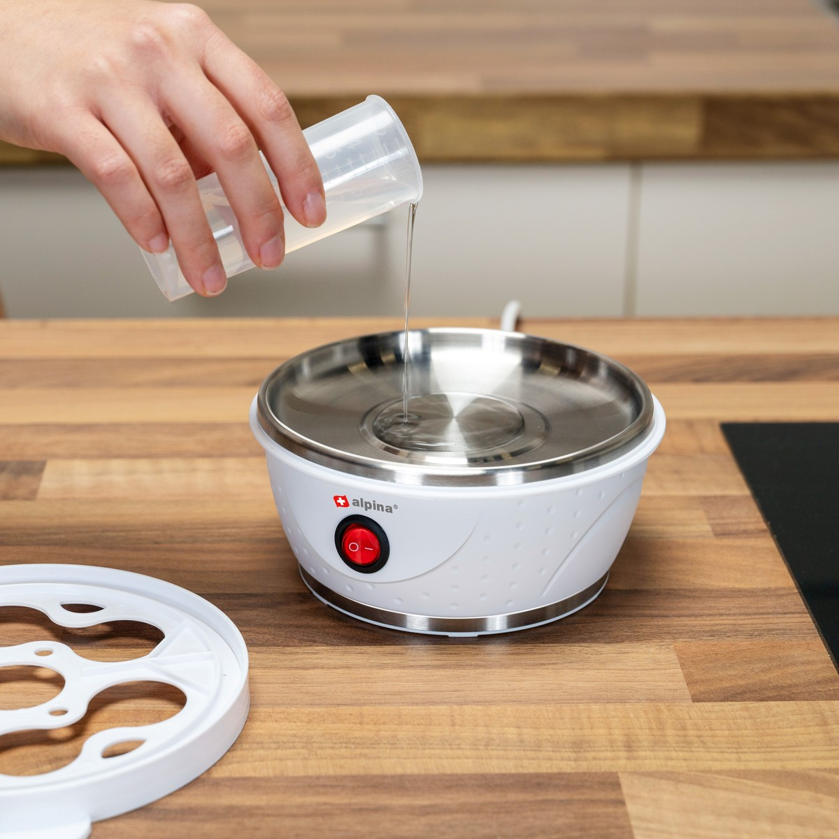 A hand pours water from a measuring cup into a white egg cooker on a wooden table.