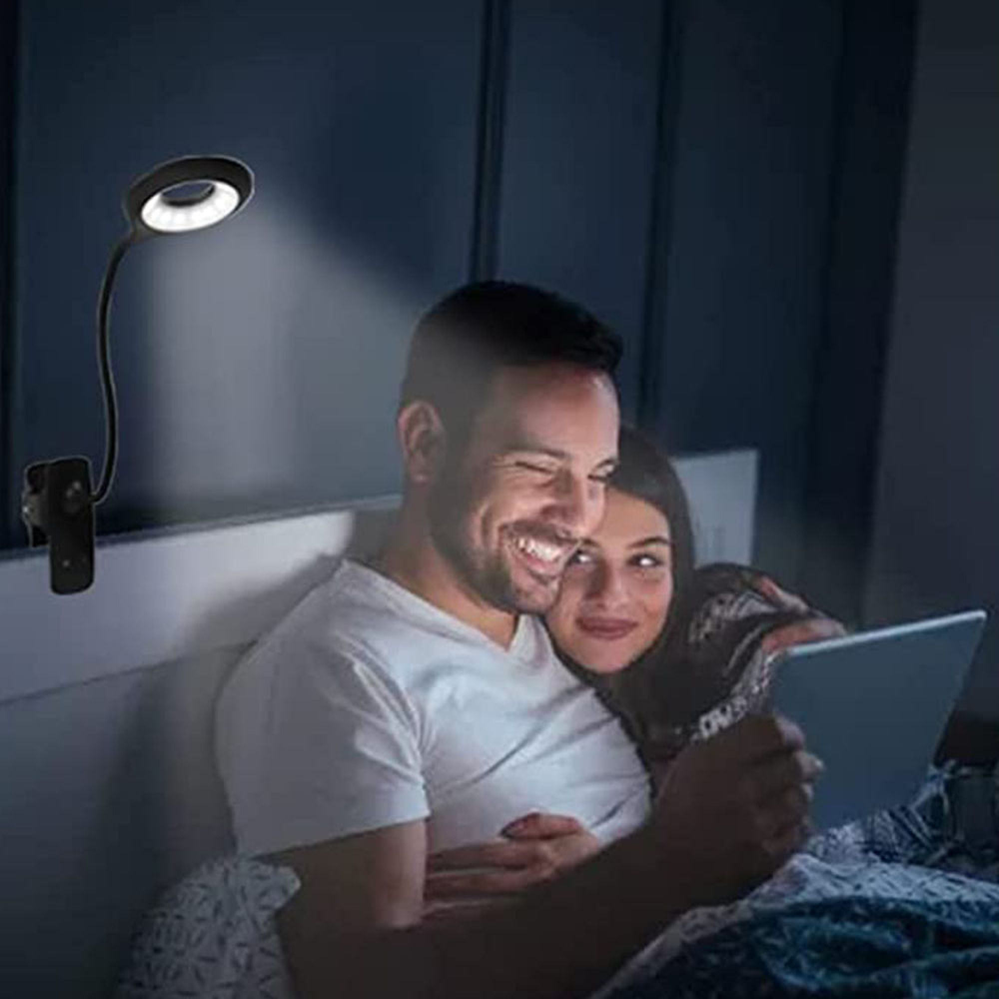 Ein Paar liegt im Bett. Eine Klemm-Ringlampe beleuchtet sie, während sie ein Tablet nutzen.