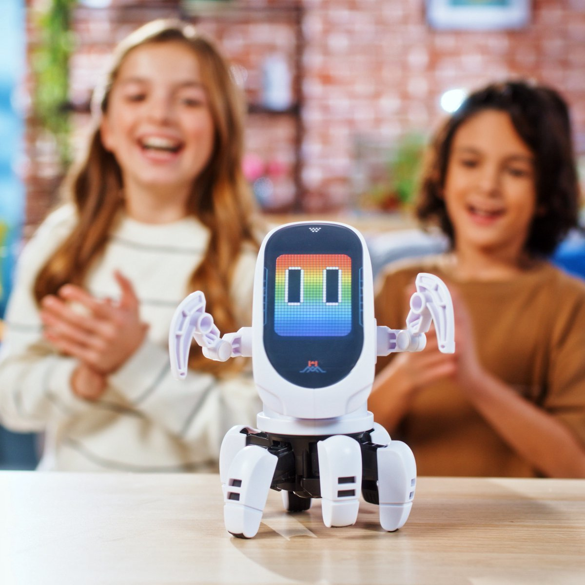 Un piccolo robot con occhi colorati su un tavolo, bambini che applaudono sullo sfondo.