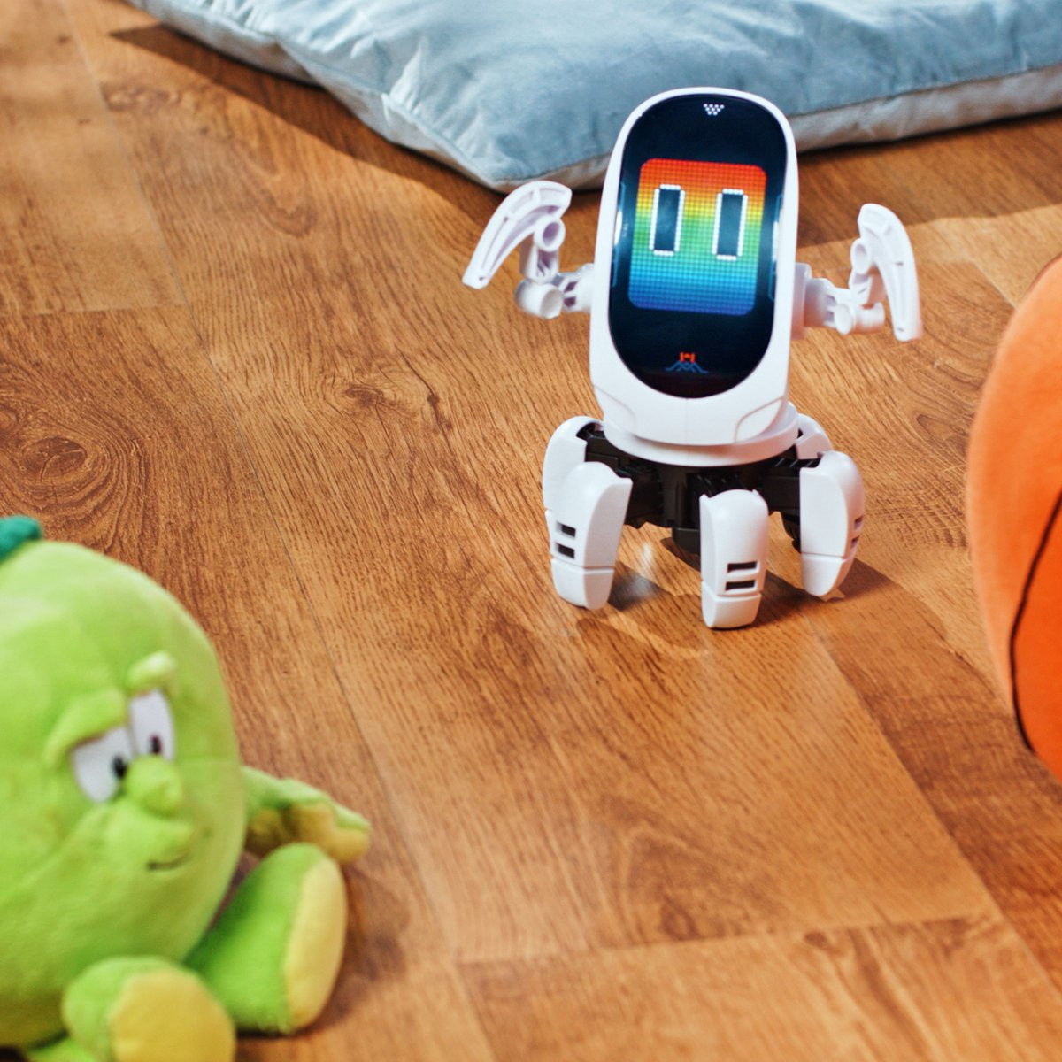 Un robot bianco con schermo arcobaleno sta su un pavimento di legno accanto a un peluche verde.