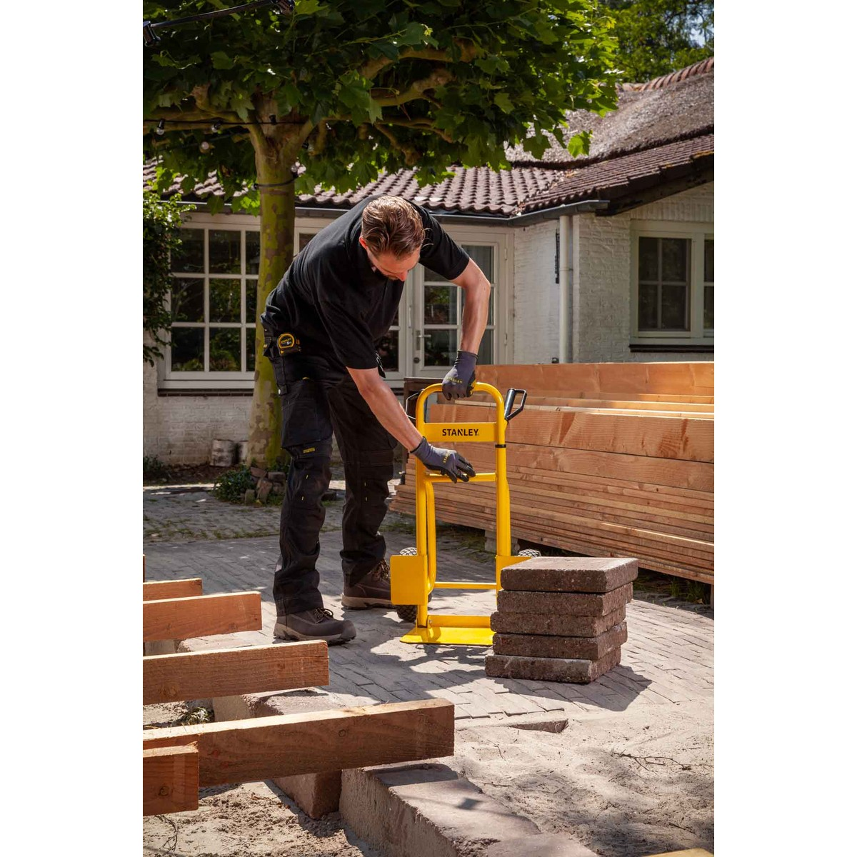 Man loads yellow hand truck with bricks in front of wood stacks.