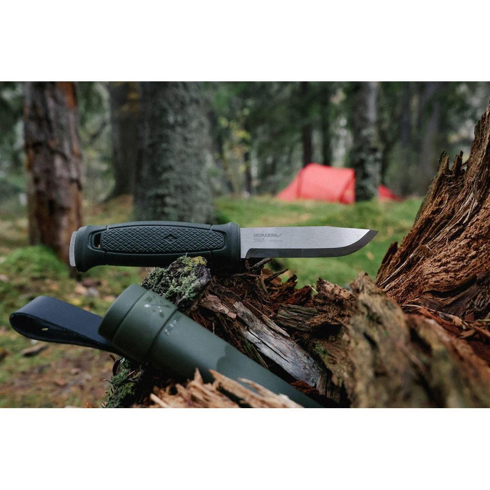 Ein Morakniv-Messer und eine Scheide liegen auf einem moosbewachsenen Baumstamm im Wald mit Zelten im Hintergrund.