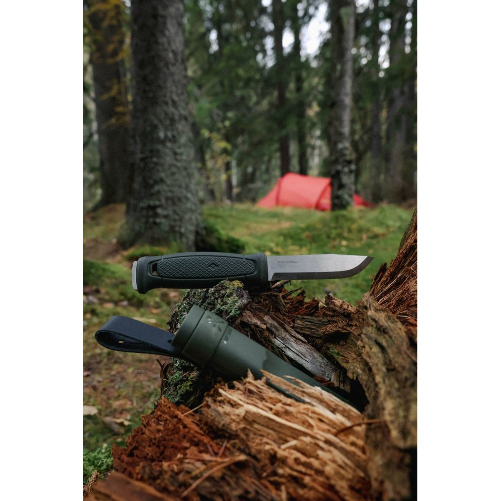 Ein Messer und seine Scheide liegen auf einem Baumstamm im Wald, ein Zelt ist im Hintergrund.