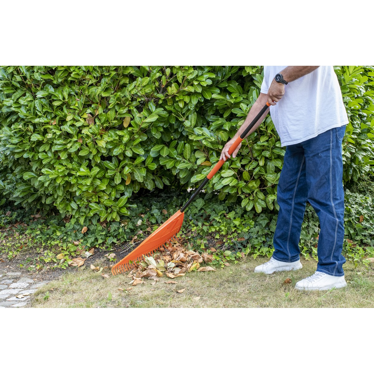 Man raking leaves in the garden with an orange rake.