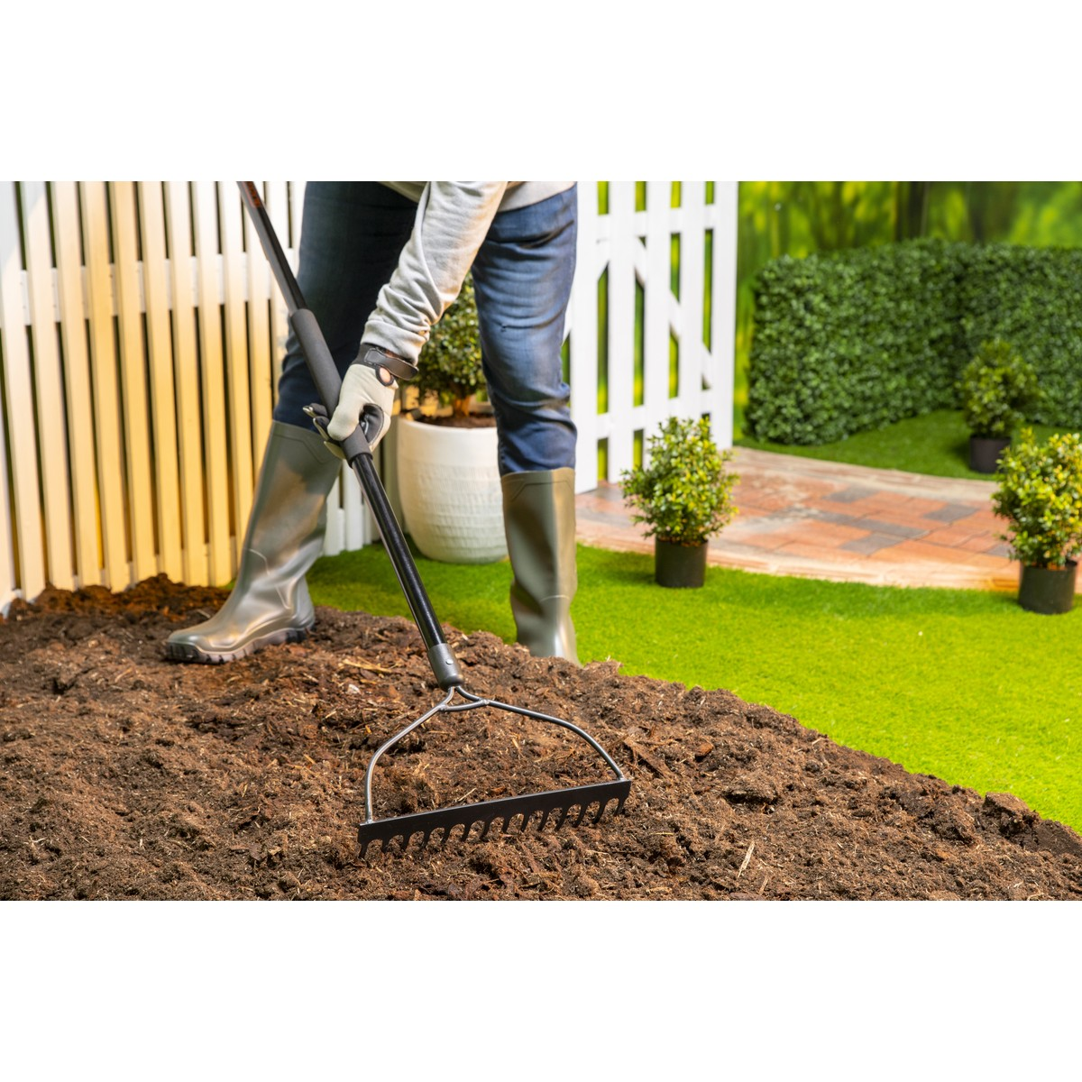 A person in boots uses a rake in dark soil near a white fence.