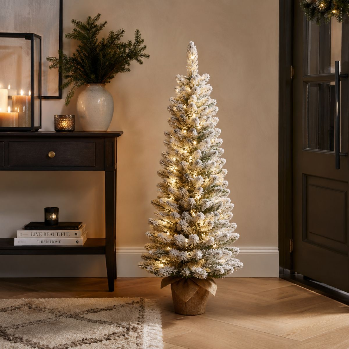 A lit artificial Christmas tree with snow effect stands next to a table with candles.