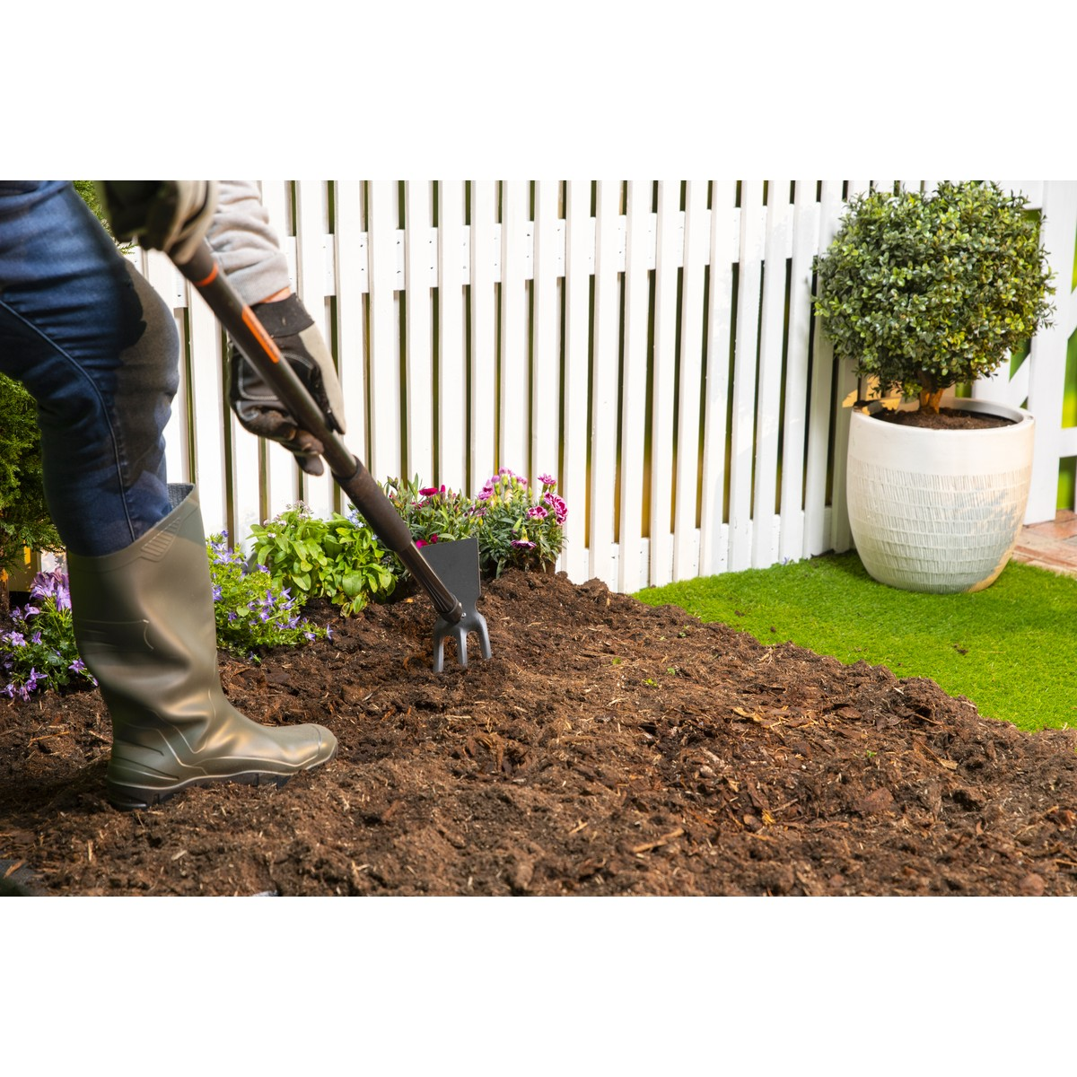Gardener tending soil with rake in front of white fence, near flowers and potted plant.