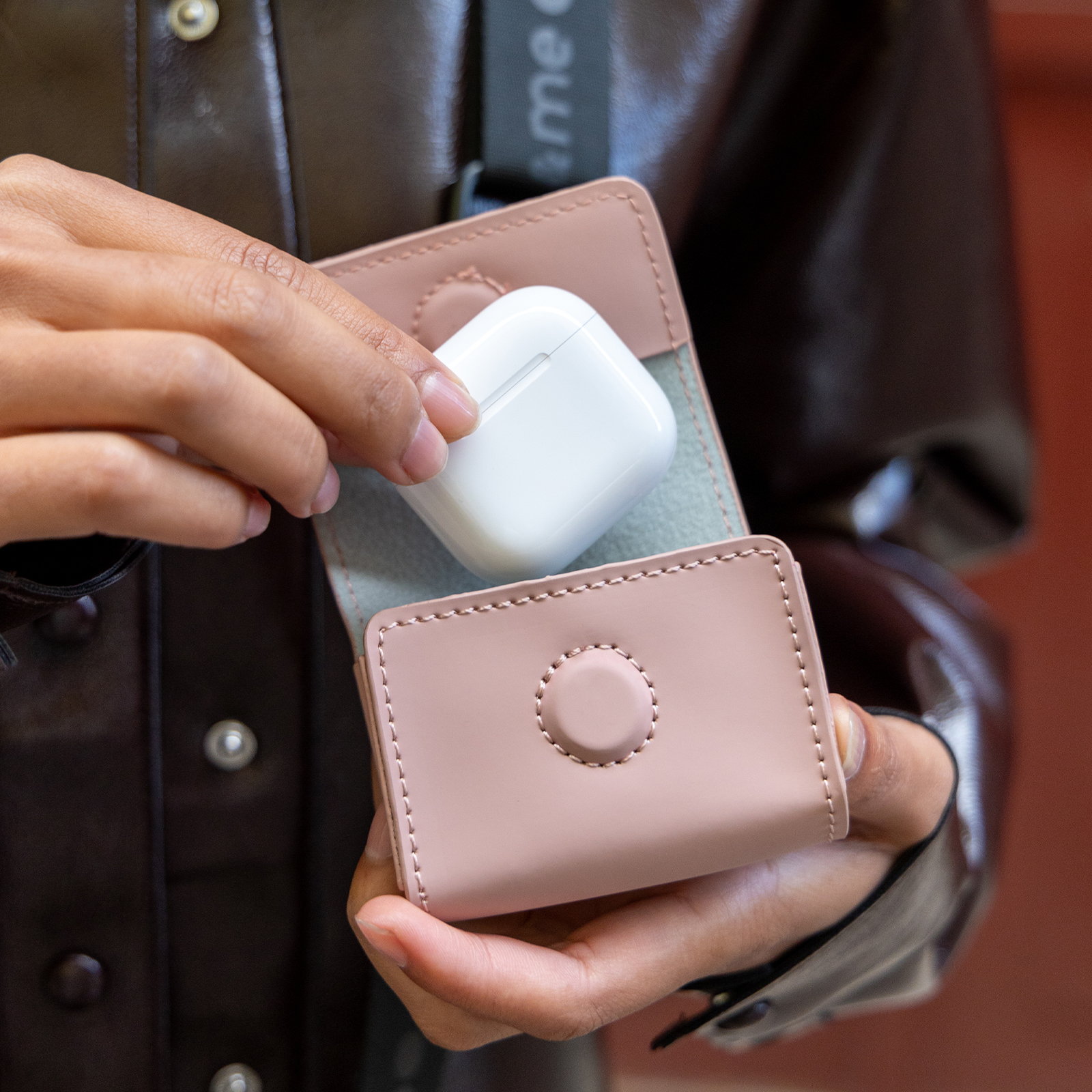 Una persona sostiene un estuche blanco para auriculares en un estuche de cuero rosa. Se ve una chaqueta marrón.