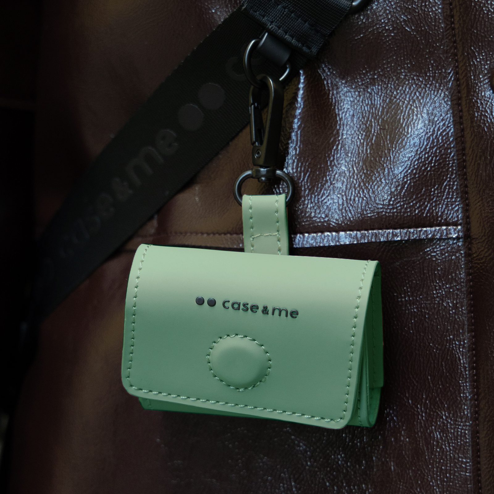 Un estuche verde claro con el logo "case & me" cuelga de una correa oscura.