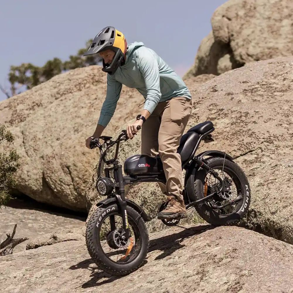 Uomo con casco guida e-bike nera su terreno roccioso. Camicia blu, pantaloni beige.
