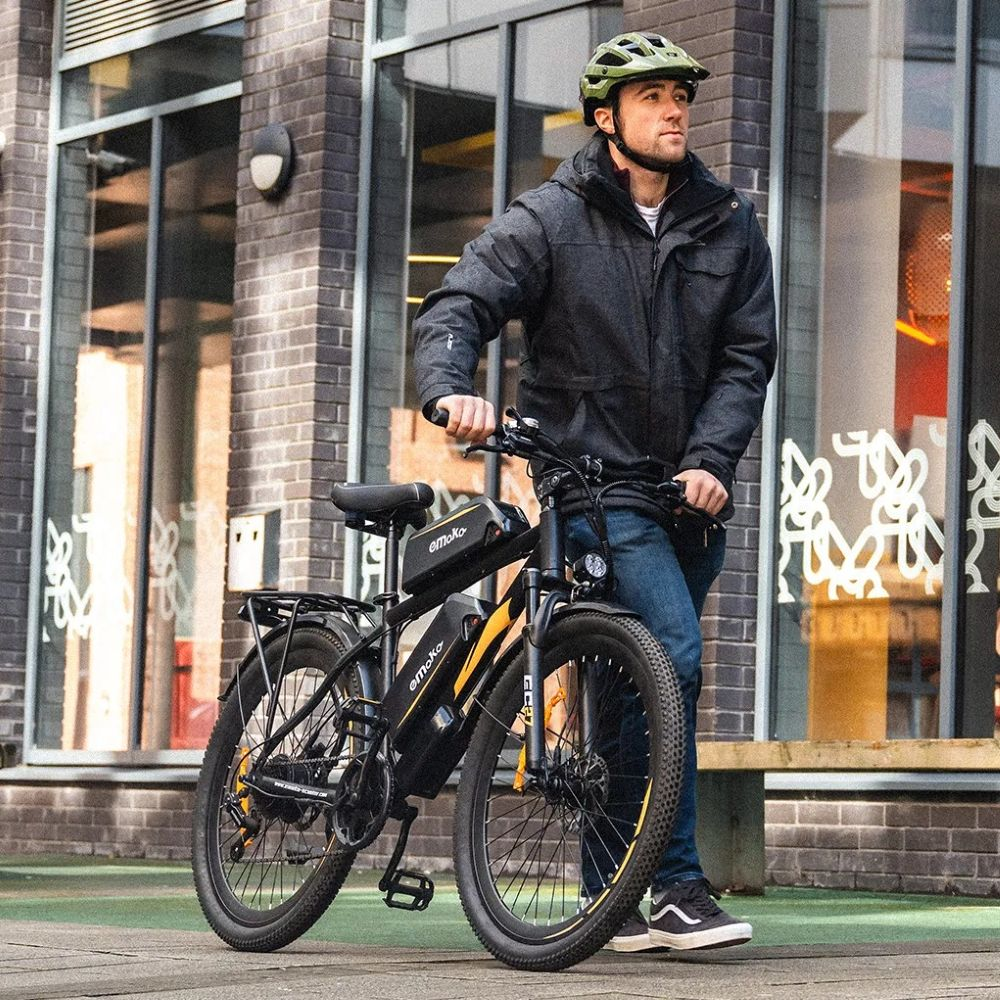 Un uomo con giacca grigia e casco è accanto a una bici elettrica nera con accenti gialli.