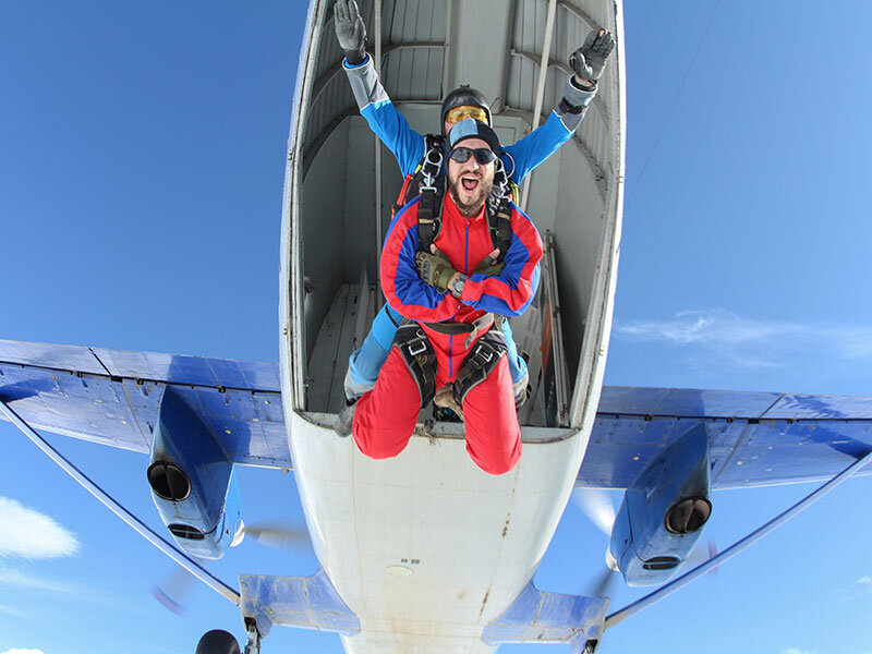 Dos paracaidistas con trajes rojos y azules saltan de un avión, en pleno vuelo, contra un cielo azul claro.